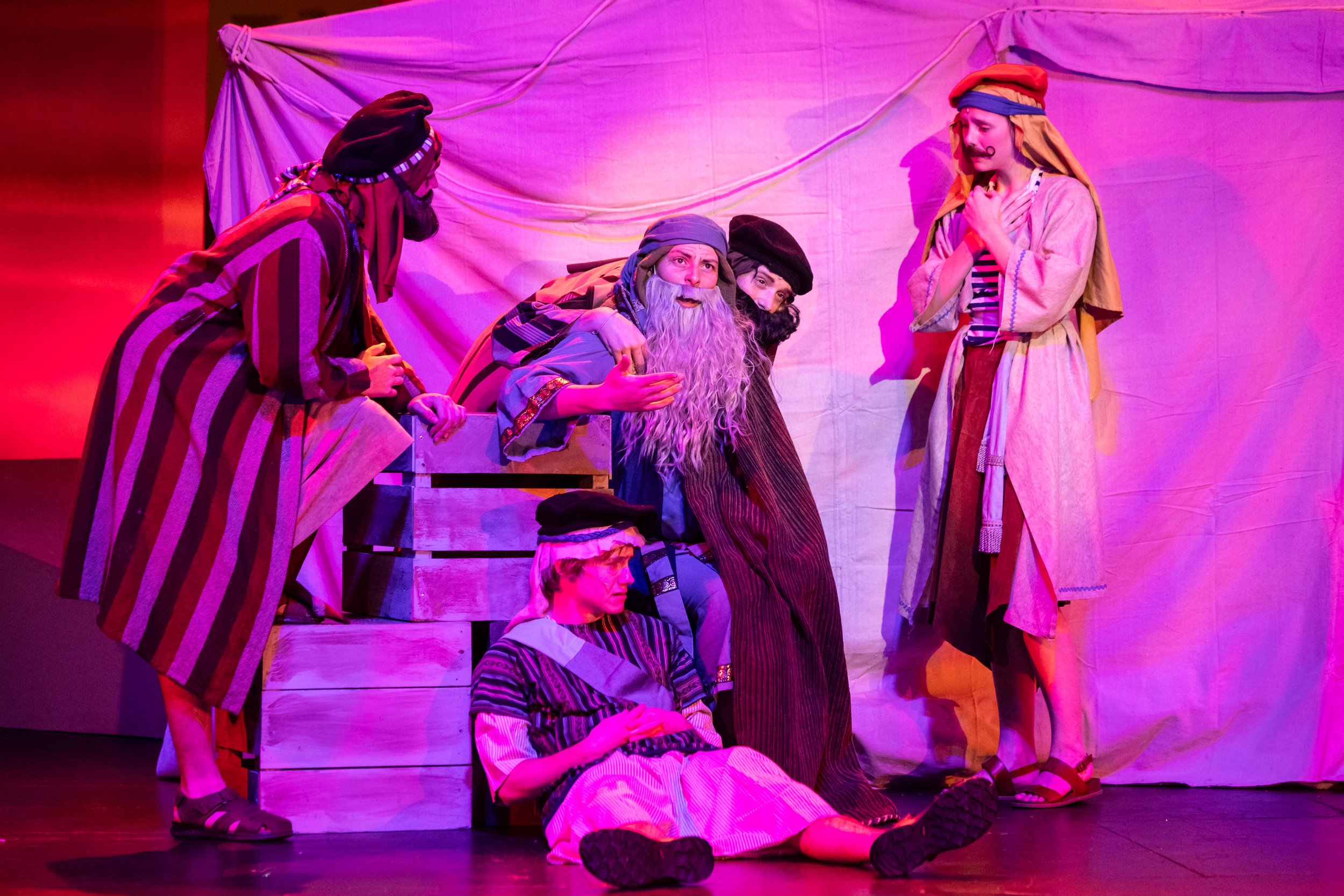 Actors performing a play on stage with colorful lighting, dressed in historical costumes, some sitting and some standing, with a white cloth backdrop.