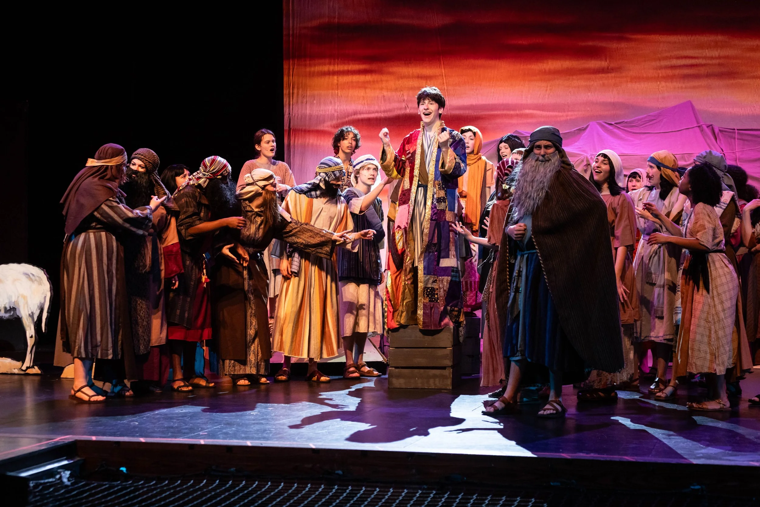A theatrical performance of a historical or biblical scene with a group of actors dressed in period costumes, on a stage with a scenic backdrop, engaged in singing or dialogue.