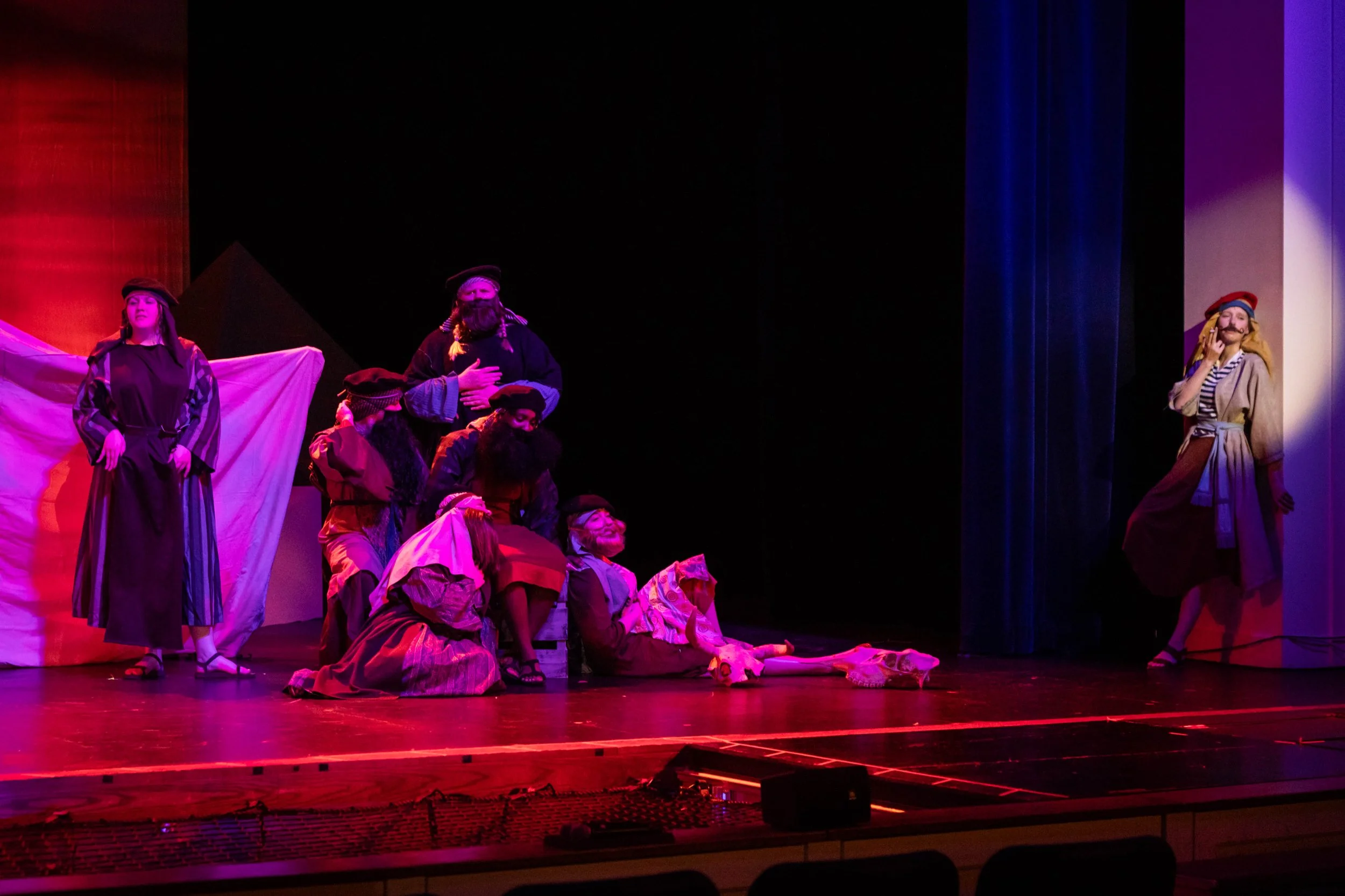 The image shows a stage with actors dressed in costumes, performing in a play with protest or historical themes, under colorful stage lighting.