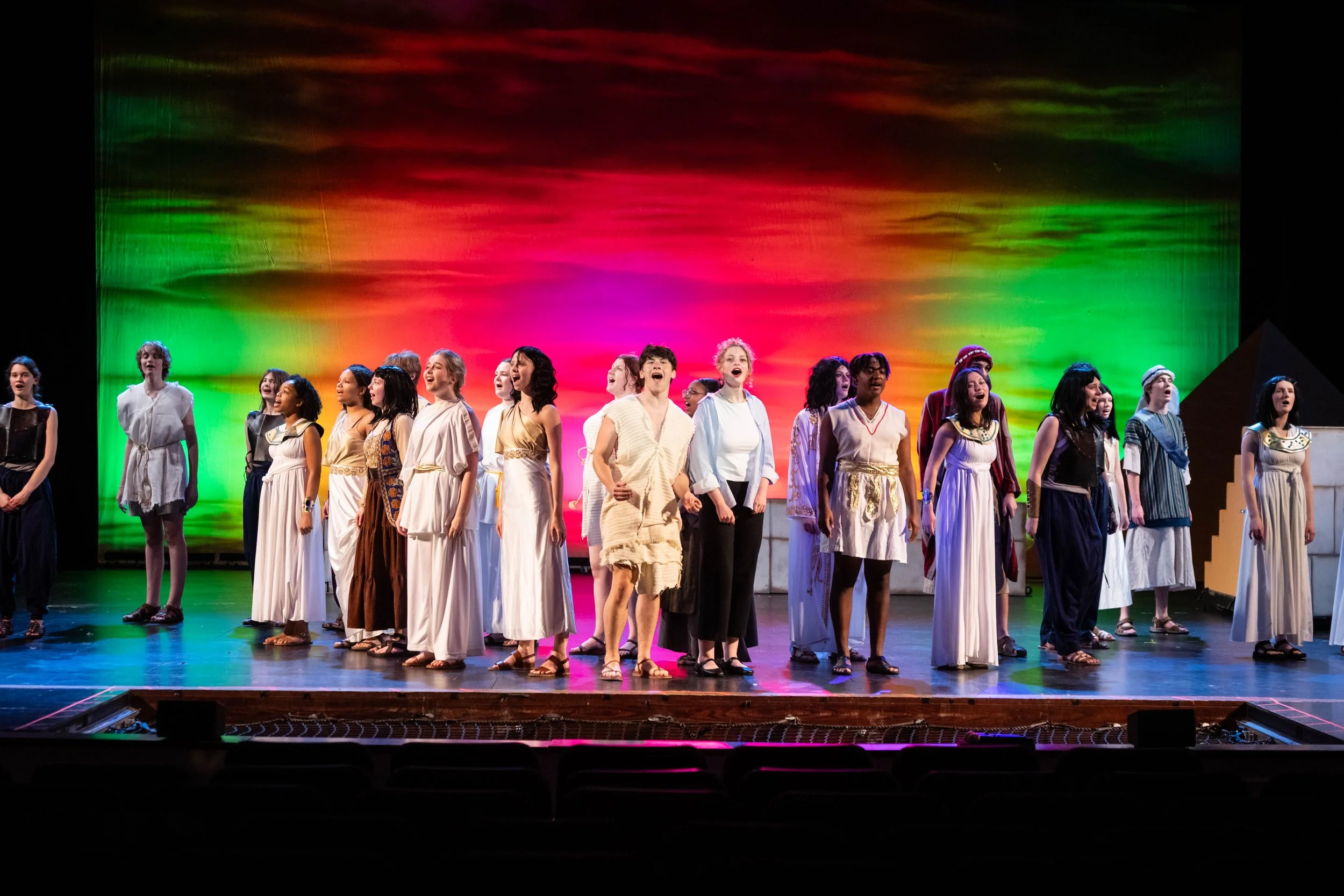 Group of diverse children and teenagers performing on stage with colorful sky backdrop.