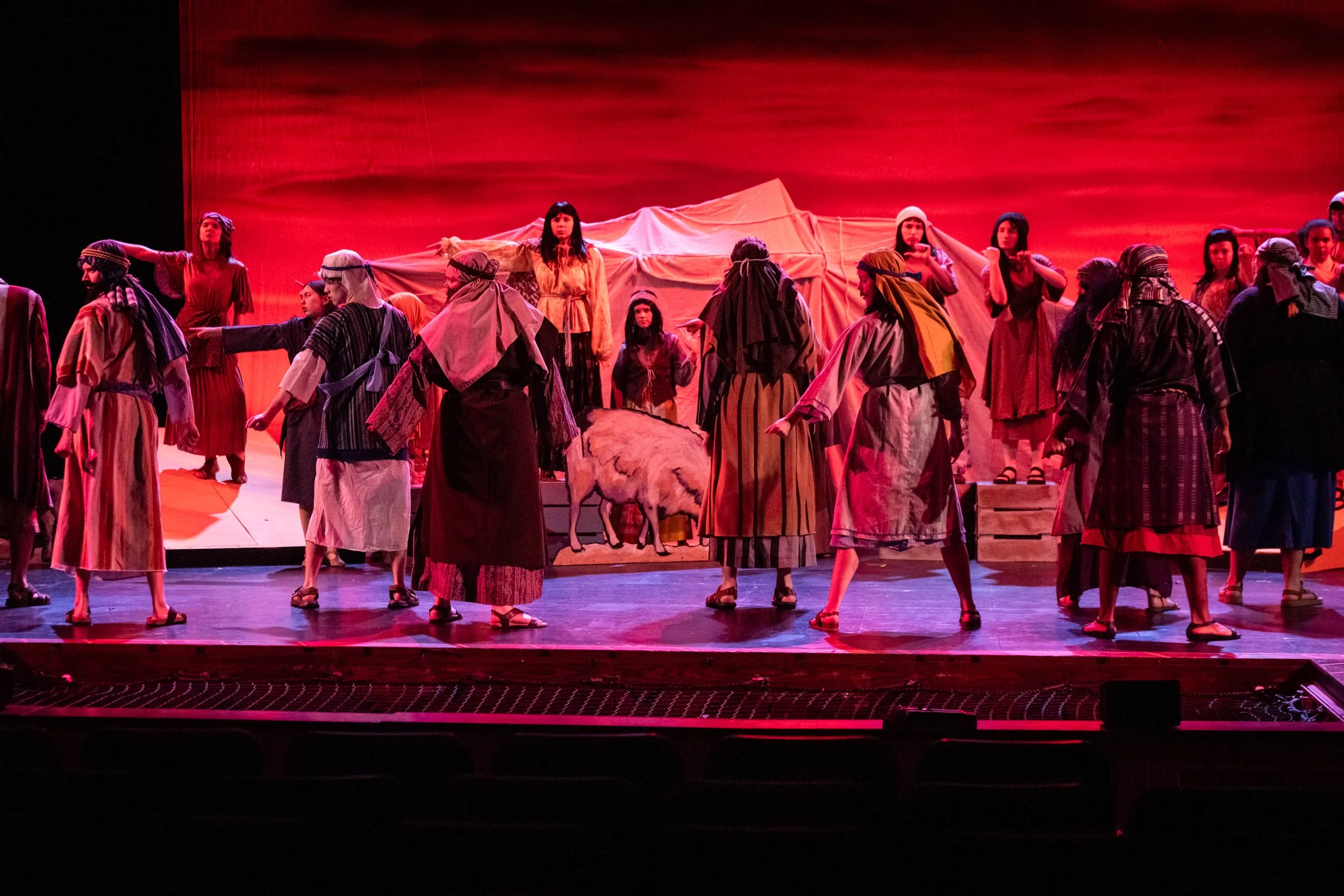 Theater scene with actors in costumes on stage, set resembling biblical nativity scene with figures dressed as shepherds and Mary, with a camel prop, under red lighting.