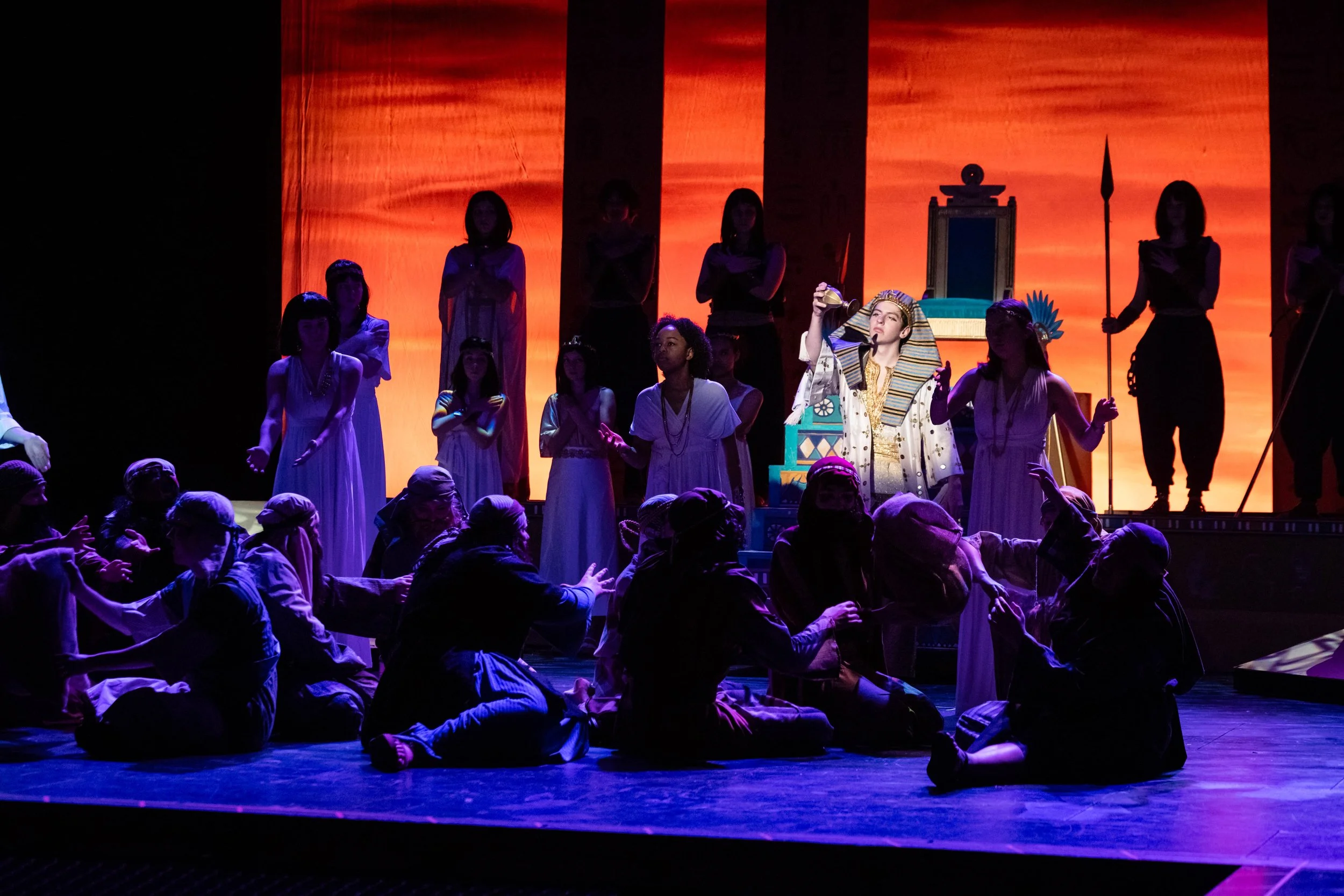 A stage performance featuring actors dressed as ancient Egyptians, with a backdrop depicting a desert scene with pyramids, and a mixture of standing and seated performers.