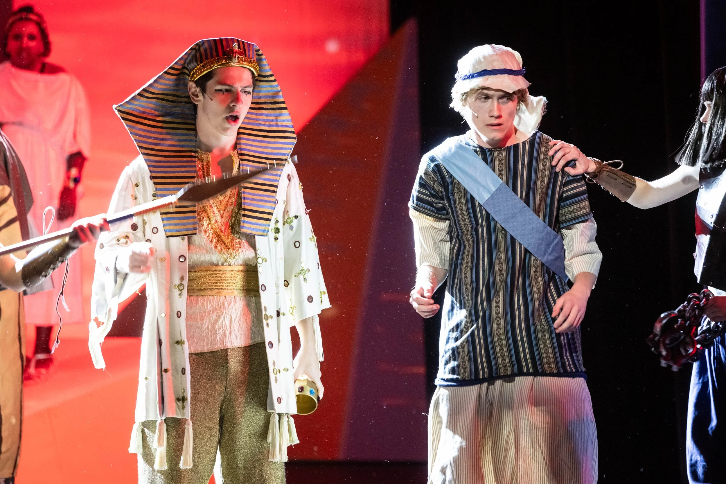 Two young actors performing in a play, one dressed as an Egyptian in a striped headdress, and the other in casual clothing with a white hat, both appearing surprised or concerned.