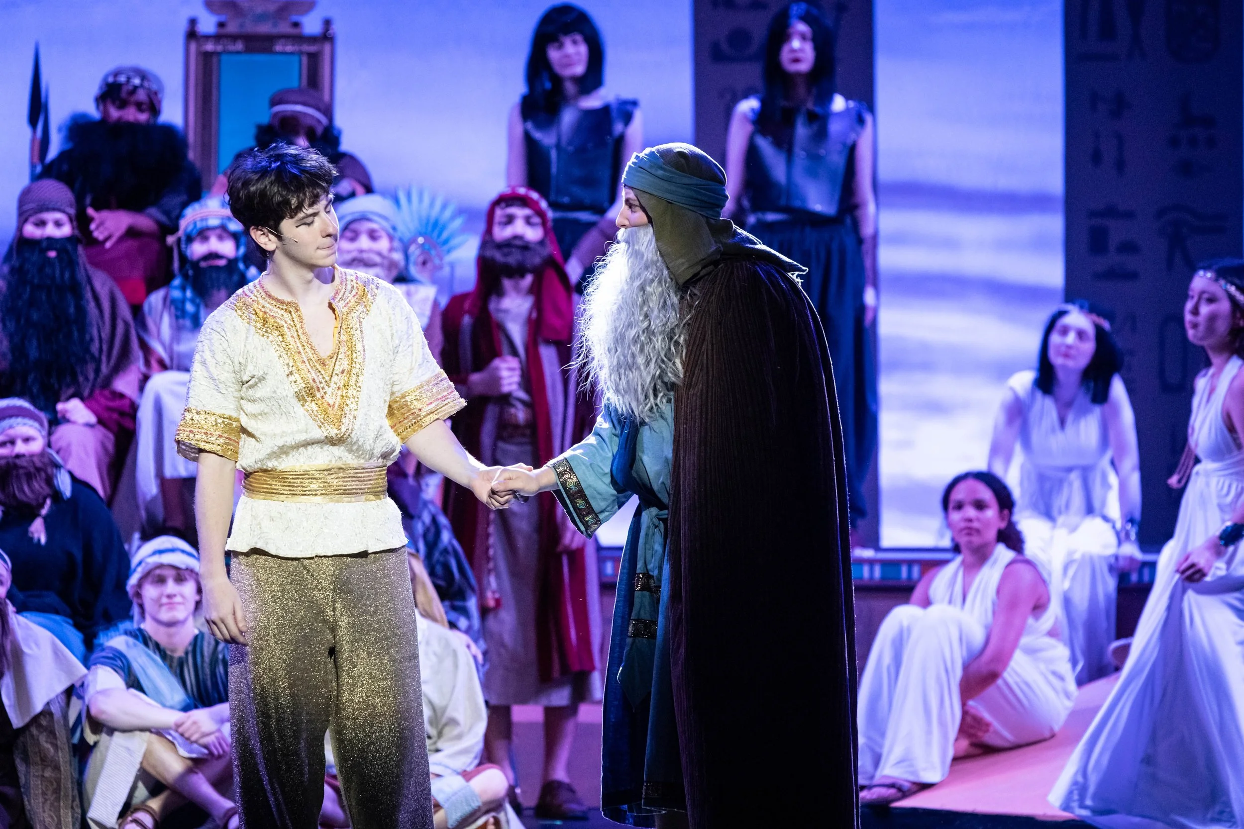 Stage scene with a woman in gold pants and a white top holding hands with a man dressed as a wizard, surrounded by performers in costumes resembling elves or dwarves.