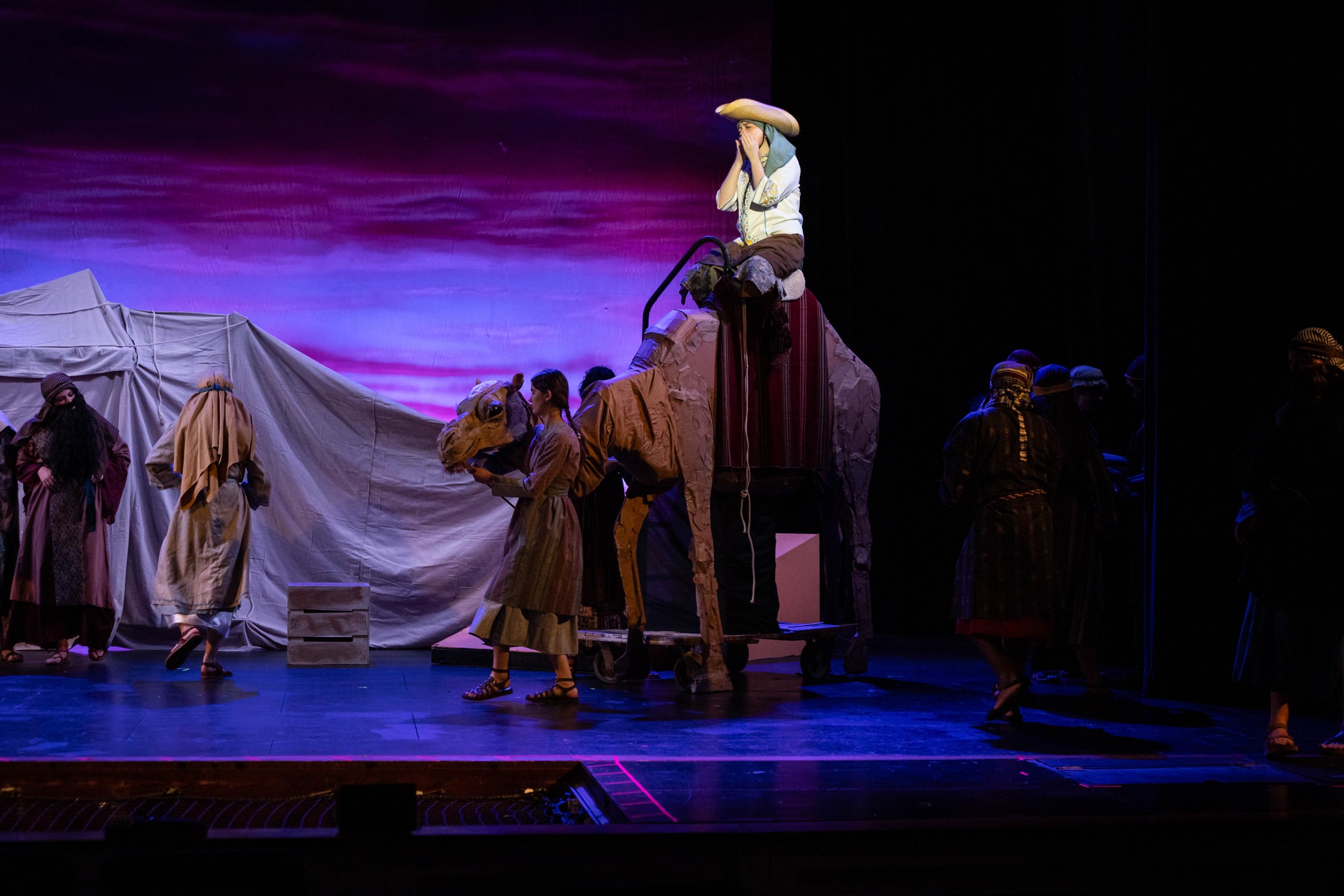 Stage scene from a theatrical performance with actors in costumes, including a woman in a straw hat, standing on a raised platform with a staircase, and other performers in period costumes, under colorful lighting.