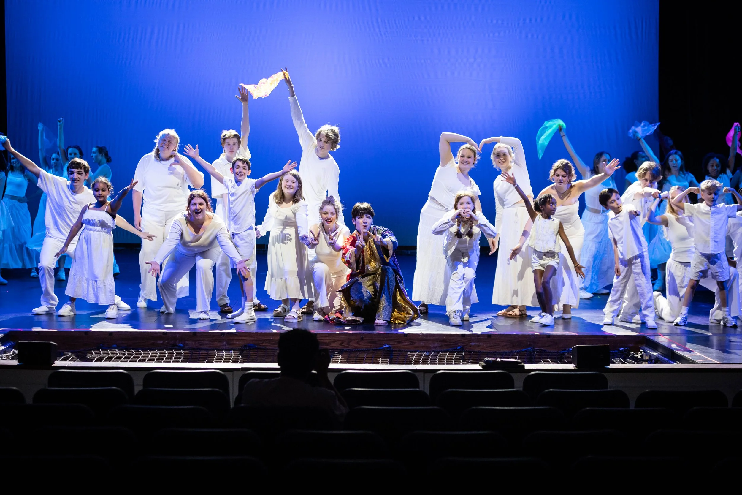 A group of children performing a dance on stage, wearing white outfits with a blue backdrop, some holding colorful scarves.