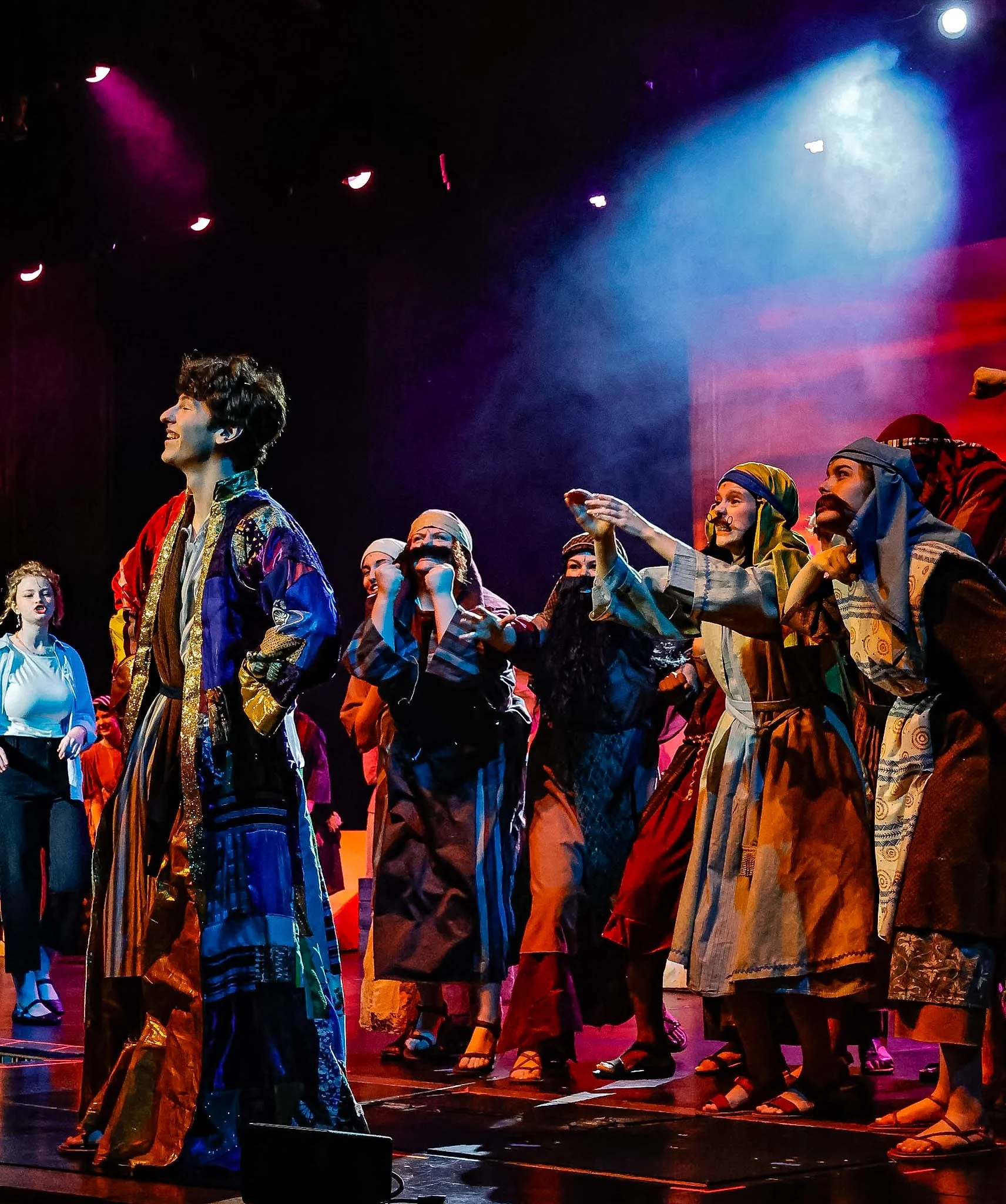 A group of performers on stage dressed in colorful costumes, engaging in a theatrical or musical performance under stage lighting, with a backdrop of vibrant colors.
