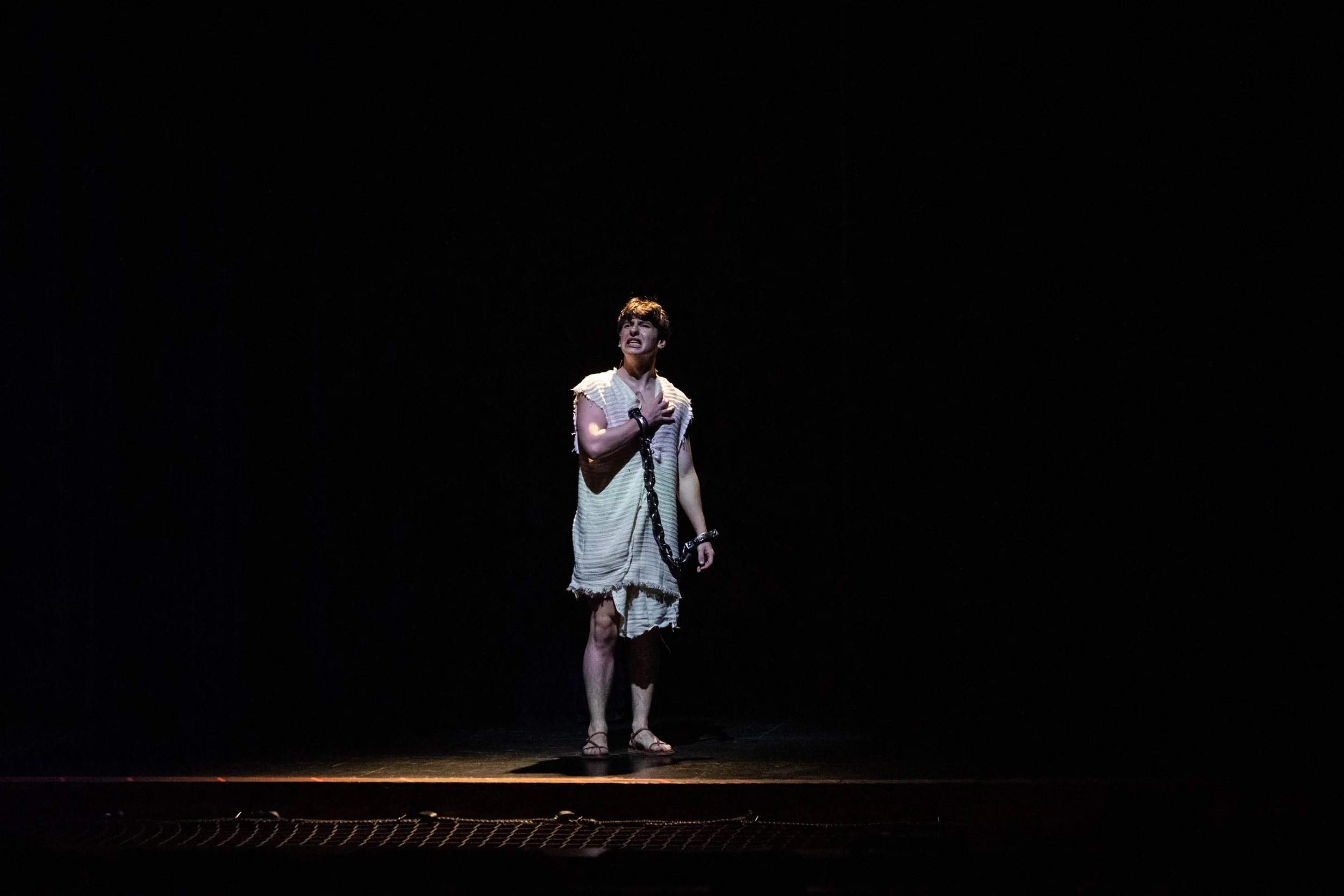 A person standing alone on stage in a spotlight with a dark background, dressed in tattered clothing, holding a chain, with a distressed expression.