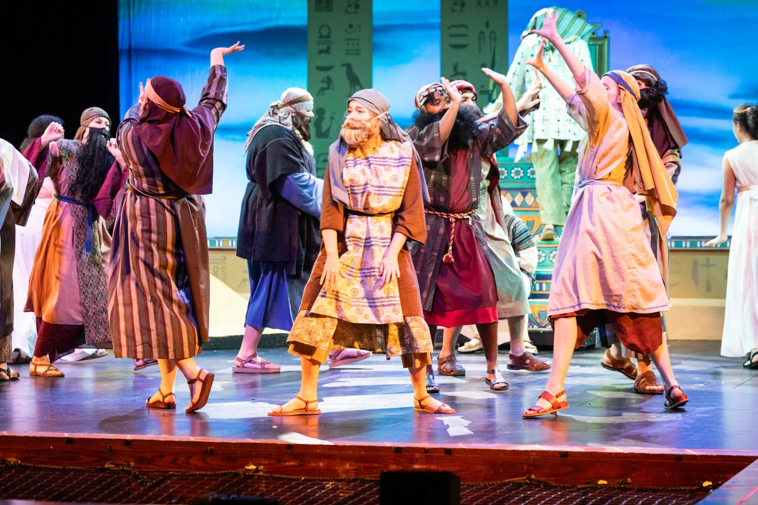 Actors dressed in biblical costumes perform on stage with a blue background and Egyptian hieroglyph-themed set pieces.