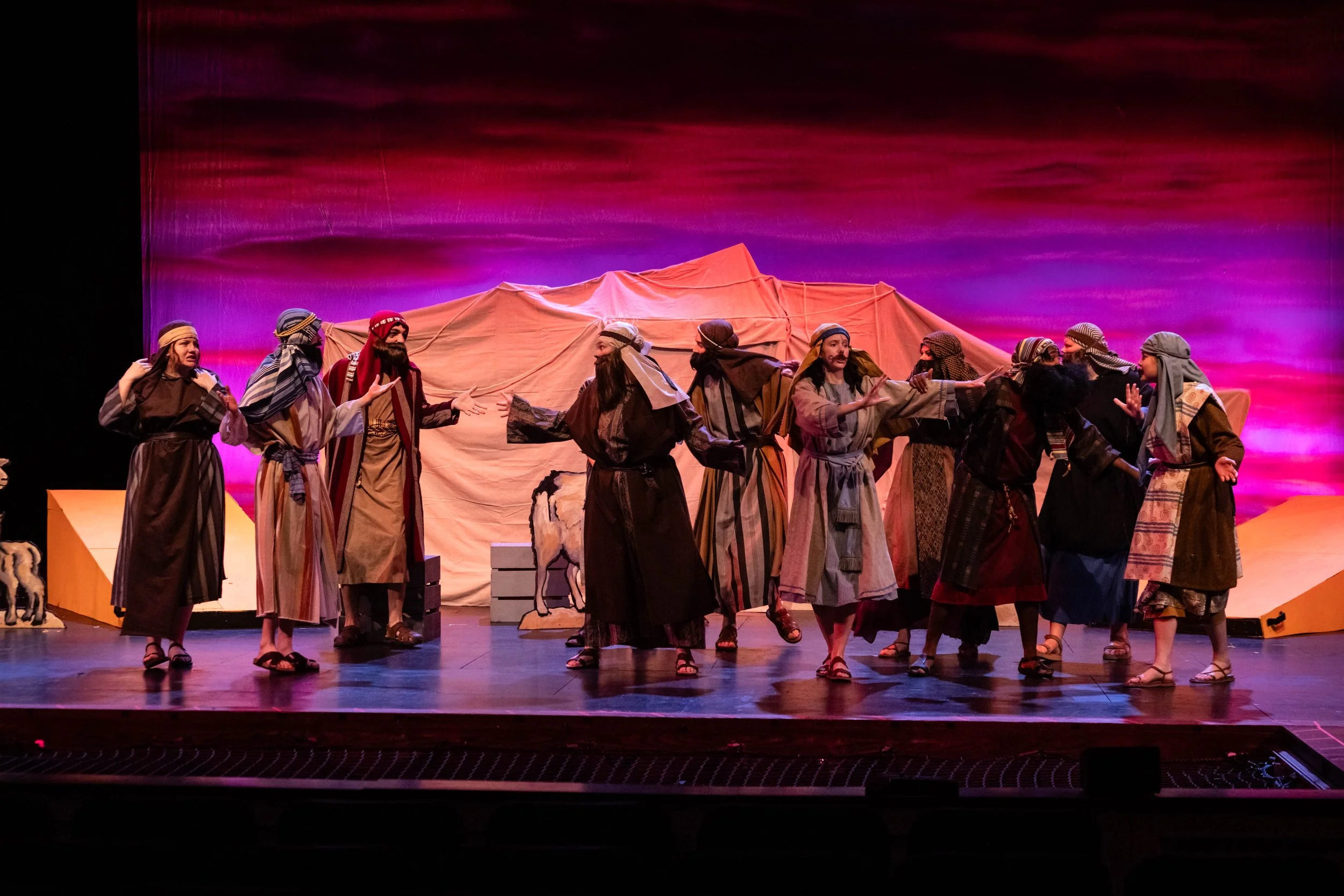 The image shows a group of actors on stage dressed in biblical costumes, performing in a play, with a colorful backdrop of pink and purple clouds.