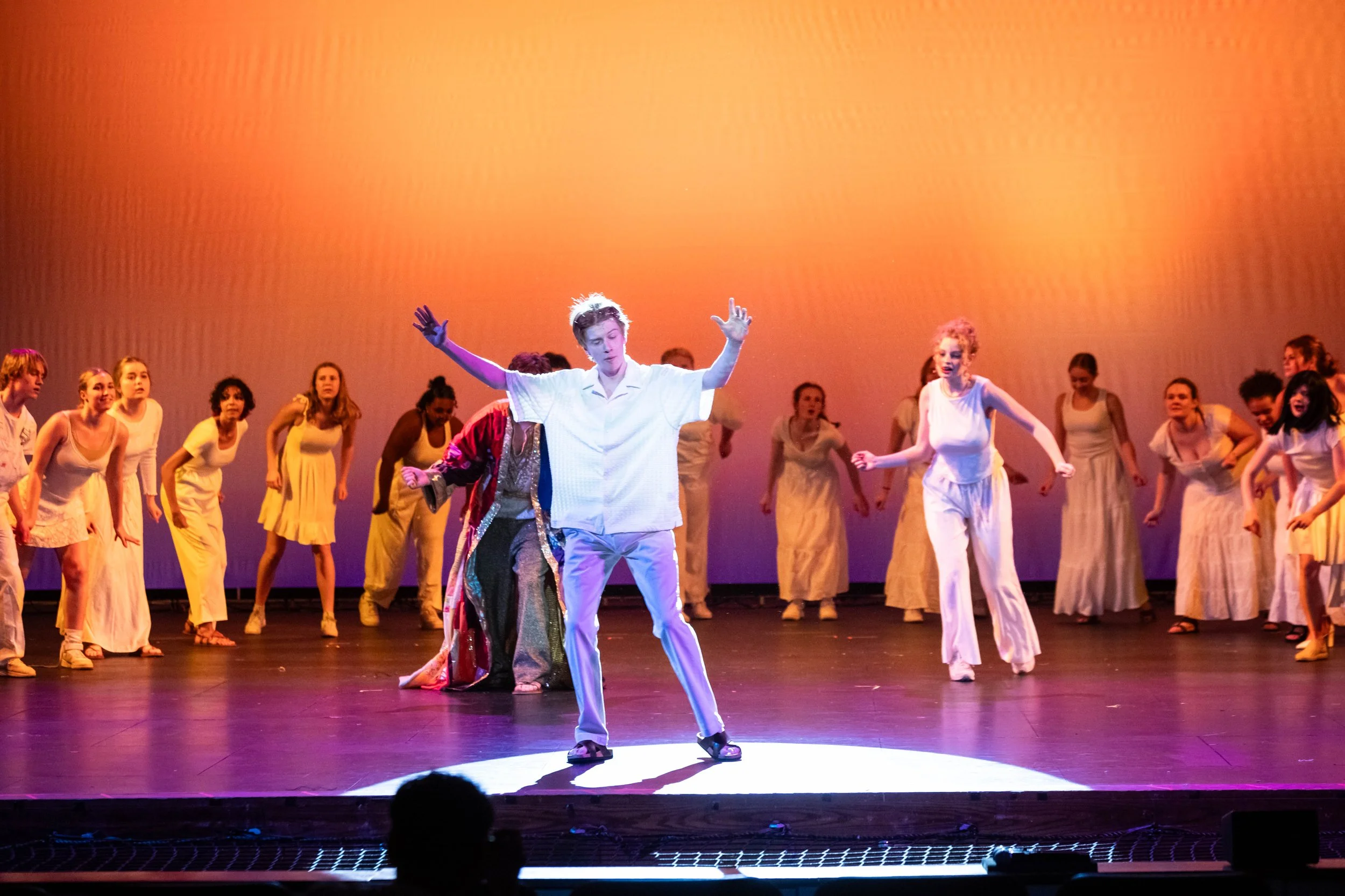 A stage performance with numerous performers, one in the center with arms raised, with a bright orange background, and other performers in white dresses surrounding him.
