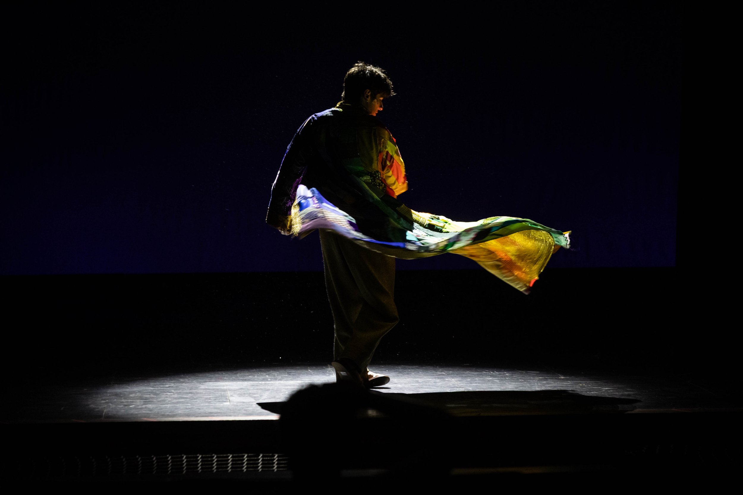 A person stands on a dark stage, illuminated from the side, holding colorful fabric, with a spotlight creating a silhouette against the black background.