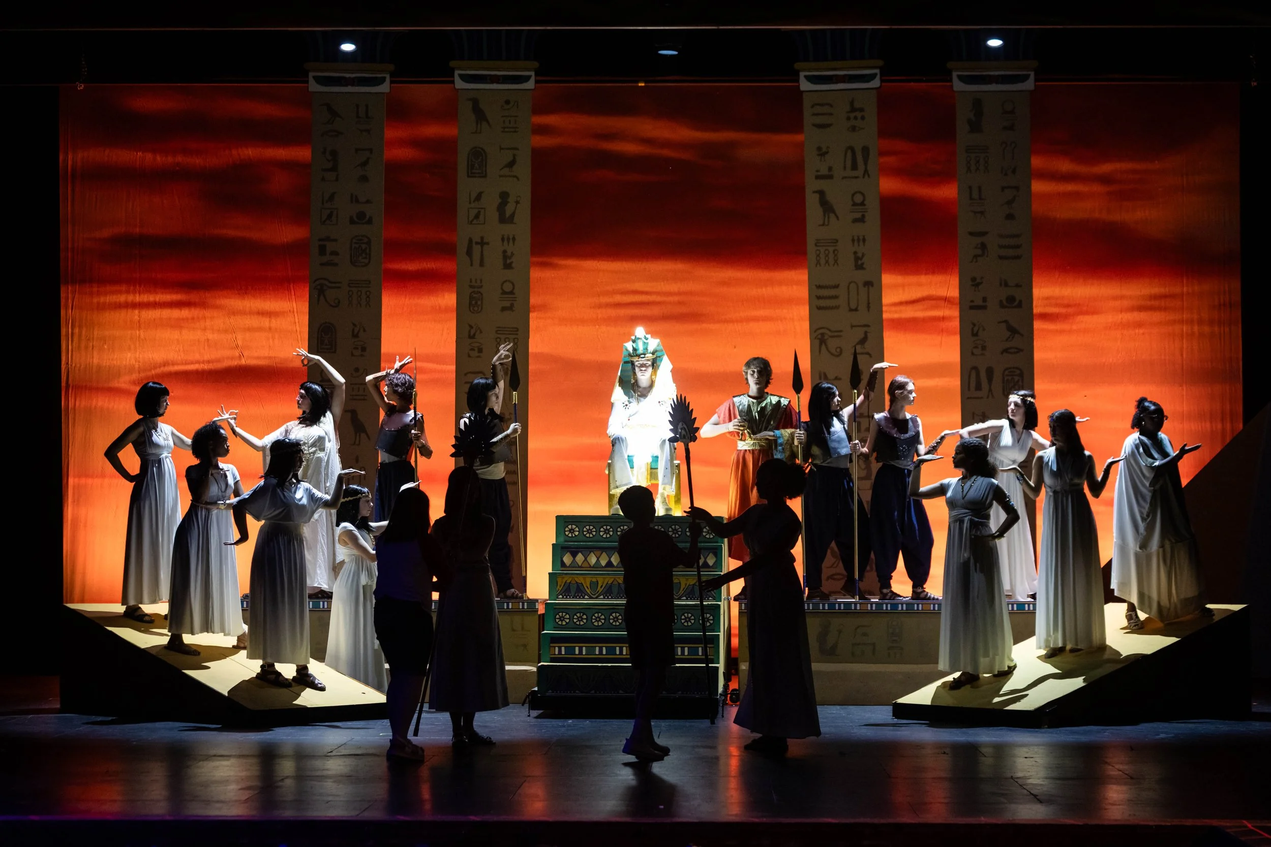 The stage performance features individuals dressed in ancient Egyptian costumes, with a backdrop resembling a sunset sky and Egyptian-themed banners hanging vertically. The scene appears to depict a historical or mythological Egyptian setting.