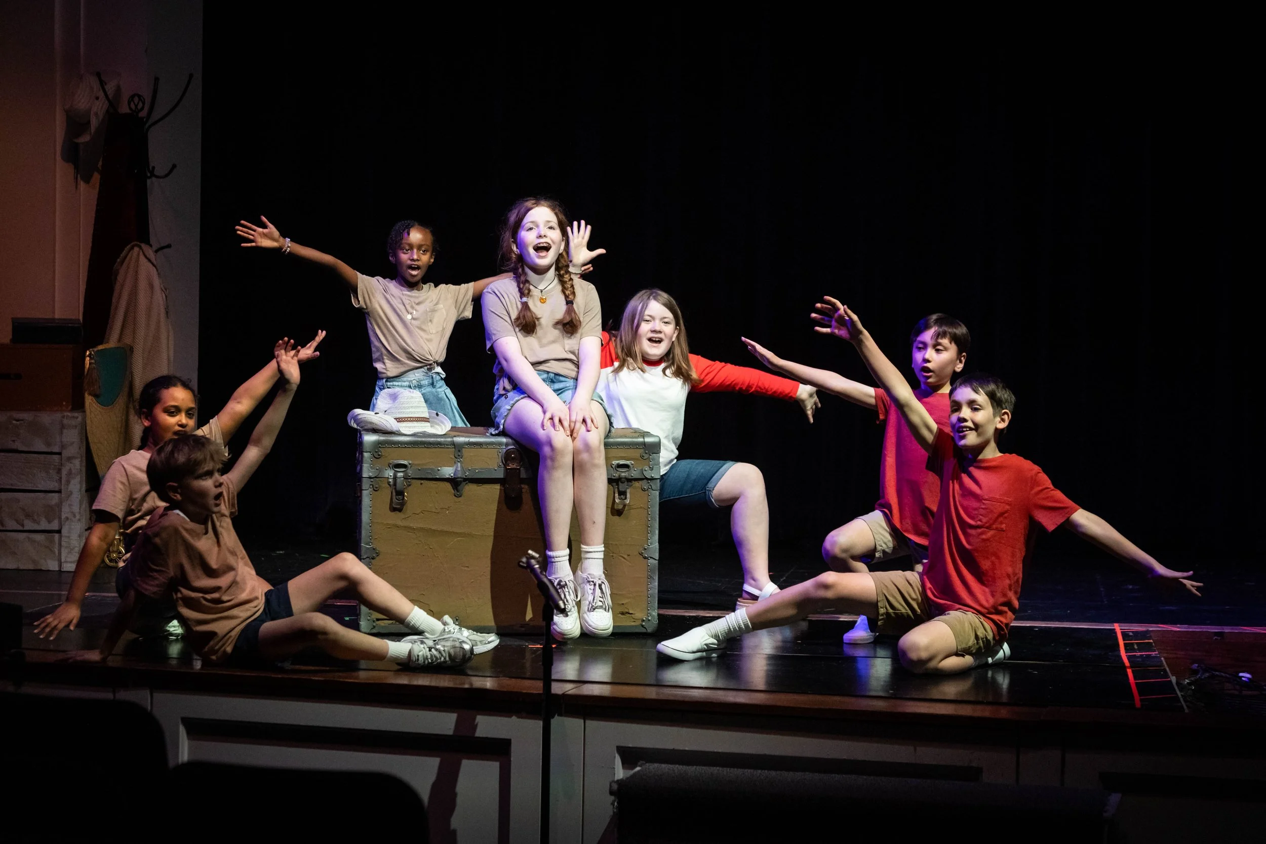 Children performing on stage during a play or musical, with some sitting and others standing, all smiling and showing expressive gestures.