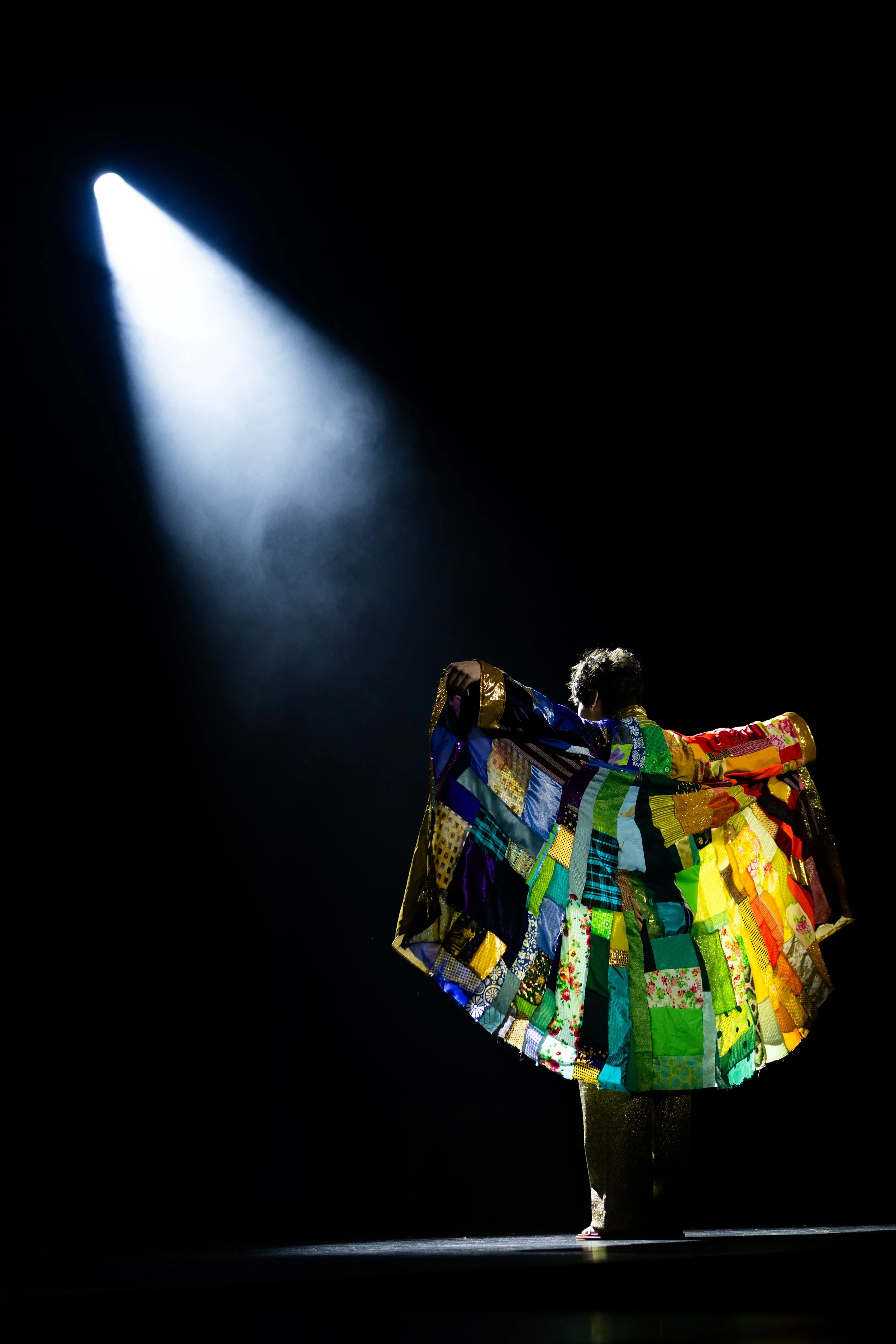 A performer on stage wears a colorful, patchwork skirt and a long sleeve top, holding out their arms as they pose in front of a spotlight against a dark background.