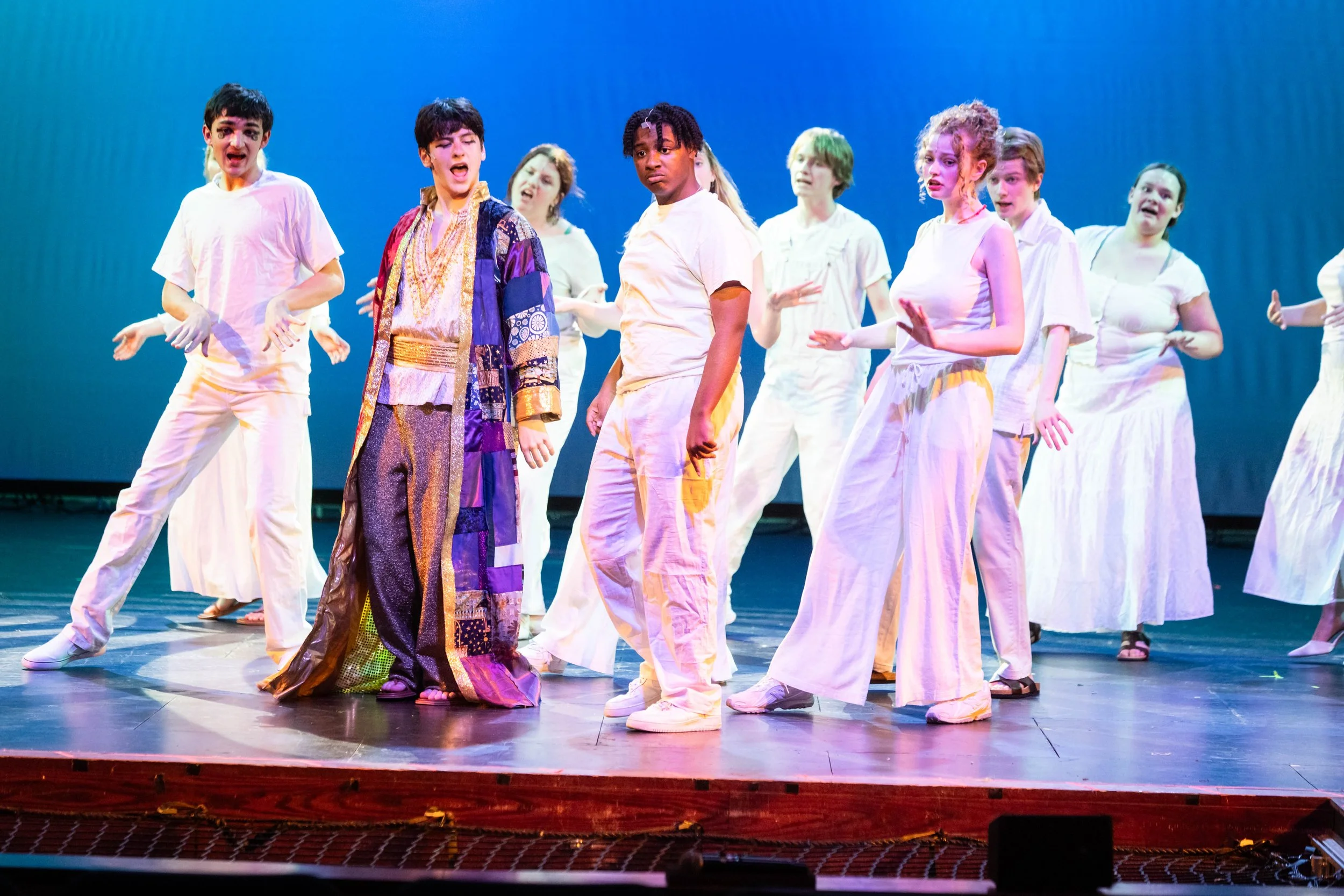 A group of young performers on stage during a theatrical or dance performance, wearing pastel-colored costumes, with some looking surprised or focused.
