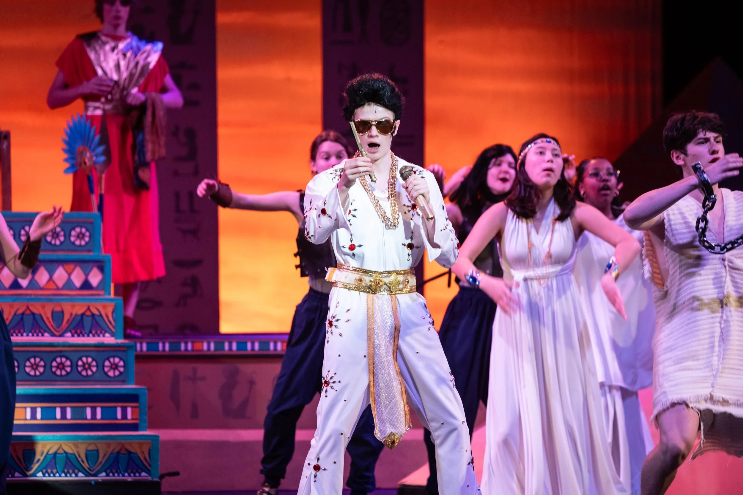A scene from a theatrical performance depicting a person dressed like Elvis Presley with sunglasses, a white jumpsuit with gold and red embroidery, holding a microphone and a gold object, surrounded by children in costumes. The background has desert 