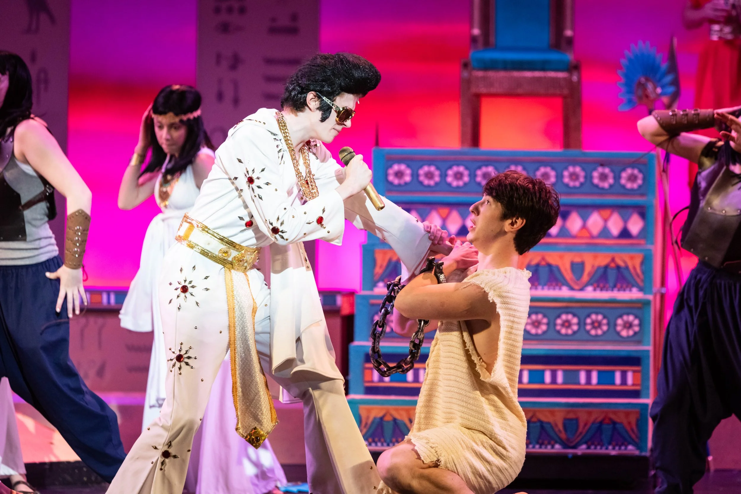 Performers on stage during a theatrical show, with one performer dressed as Elvis Presley, holding a microphone, and another actor kneeling with chains, surrounded by dancers with colorful costumes and vibrant set design.
