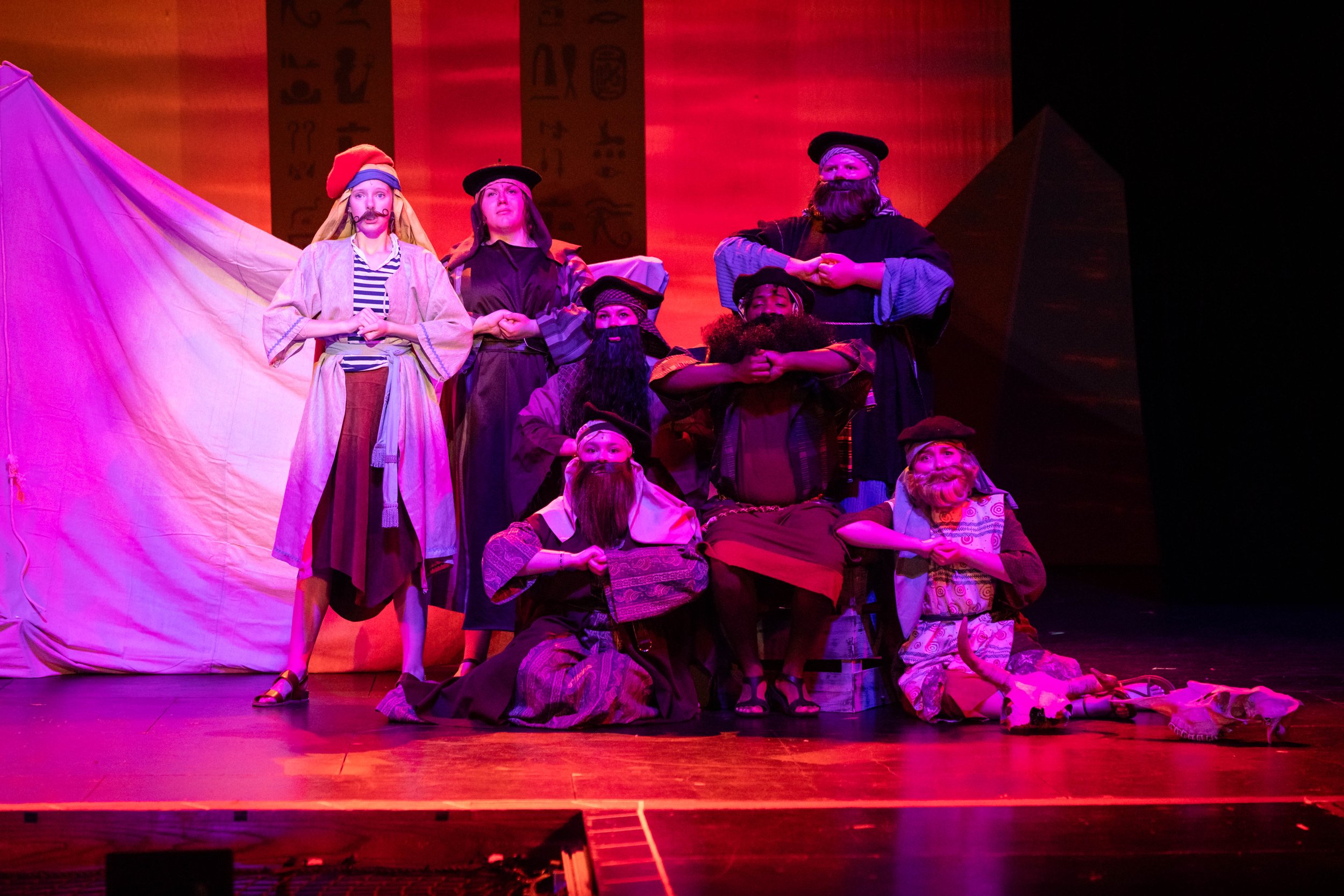 A theatrical stage with actors dressed as pirates and villagers, performing in front of a backdrop with Egyptian hieroglyphics, under pink and purple stage lighting.