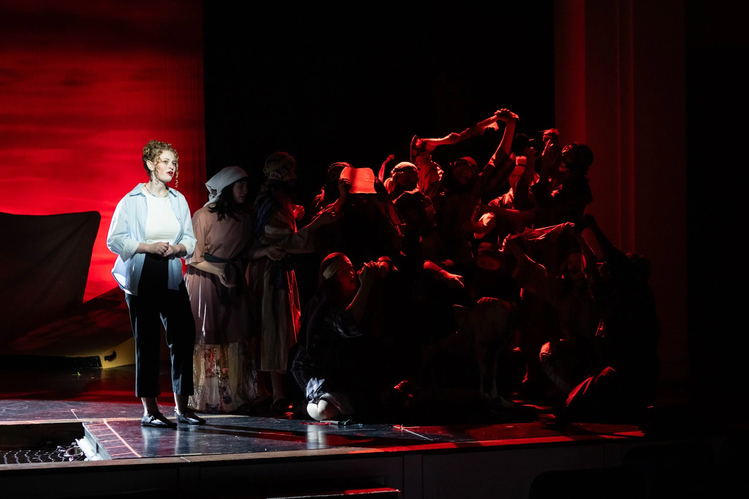 On stage, a young woman with curly hair, wearing a light blue shirt and black pants, stands alone in front of a group of performers dressed in period costumes. The group is kneeling or standing on a stairs, some raising their hands or holding props. 