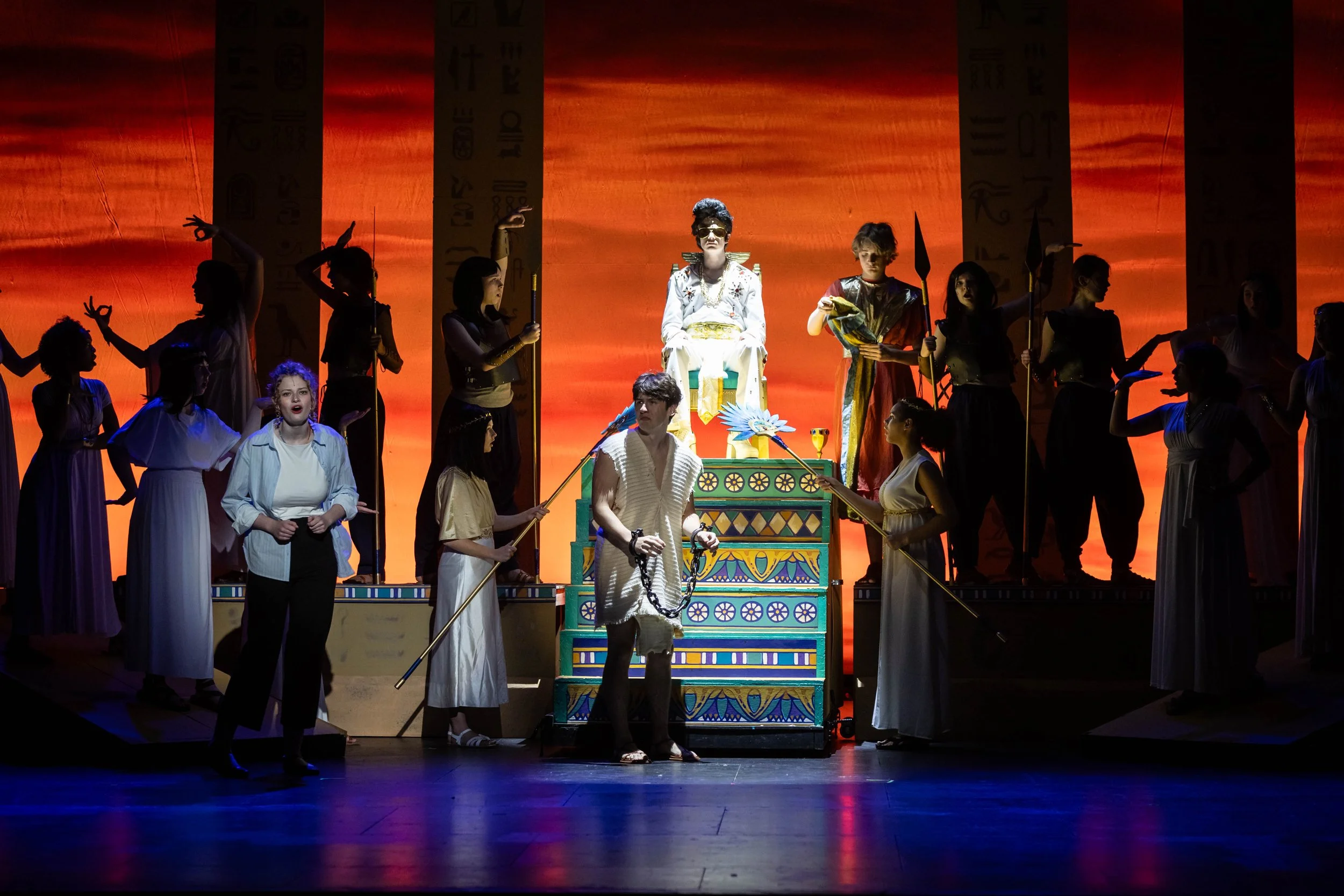 Stage scene from a theatrical performance depicting actors in ancient Egyptian costumes. The central character is seated on a throne wearing elaborate gold accessories and sunglasses, surrounded by multiple performers holding spears and other props a