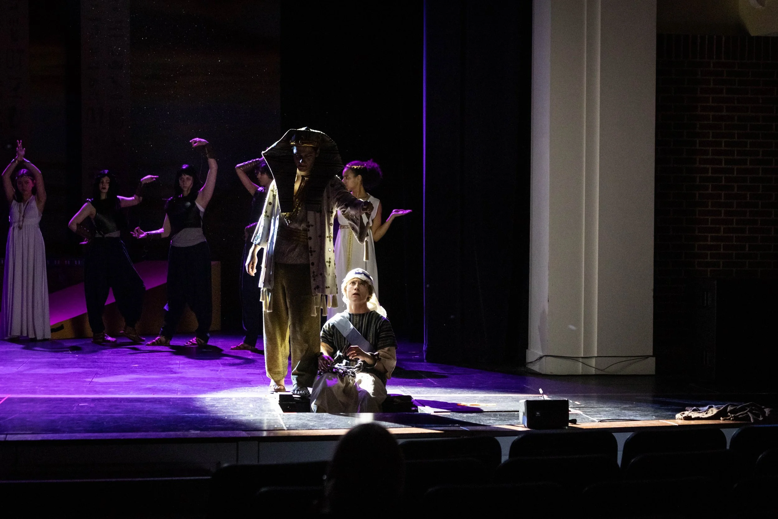 Stage performance with actors dressed in costumes, some standing and one sitting on the floor, under dim lighting with purple and yellow hues.