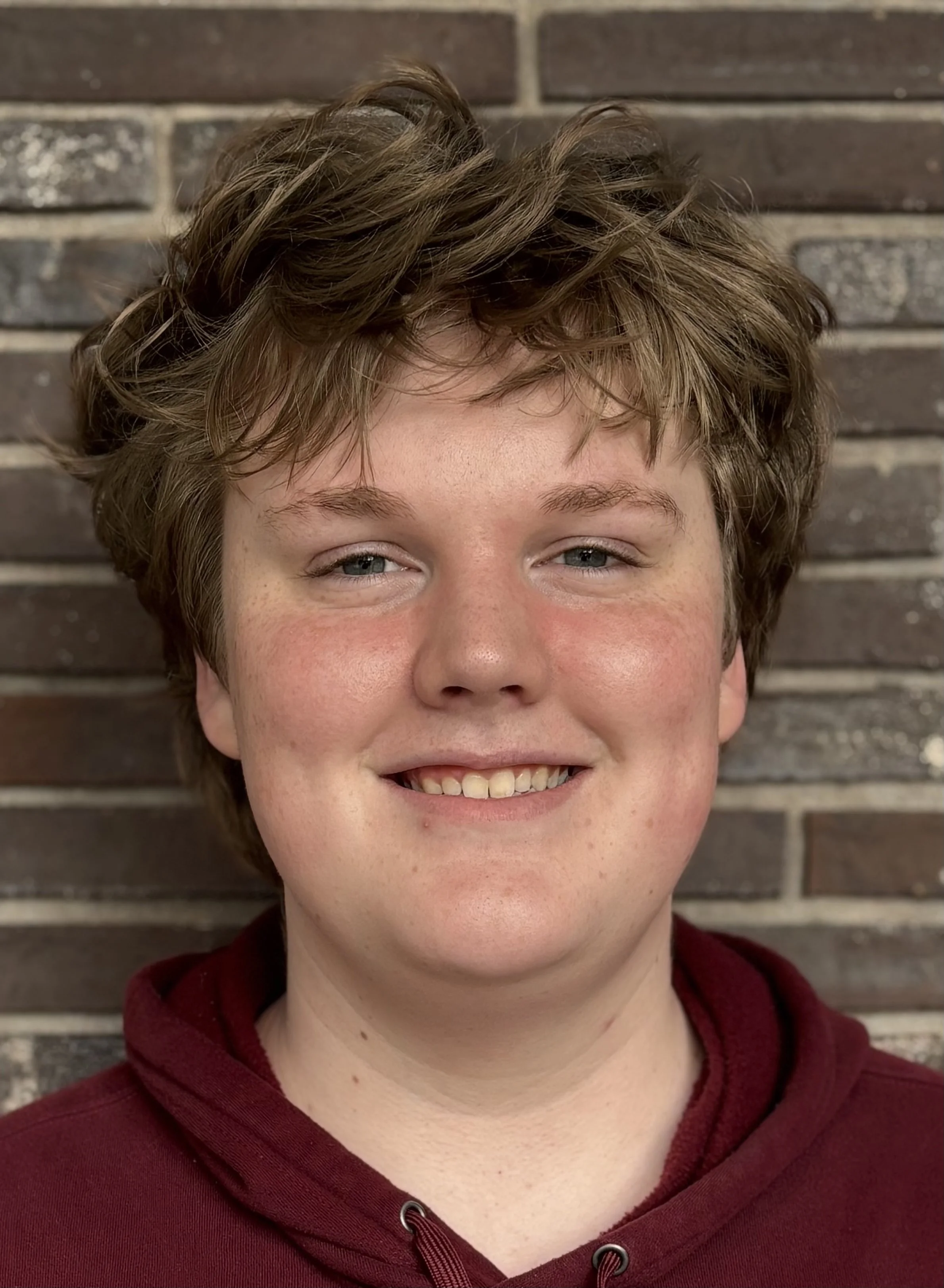 Close-up of a smiling person with curly brown hair, blue eyes, and light skin, wearing a maroon hoodie, standing against a brick wall background.