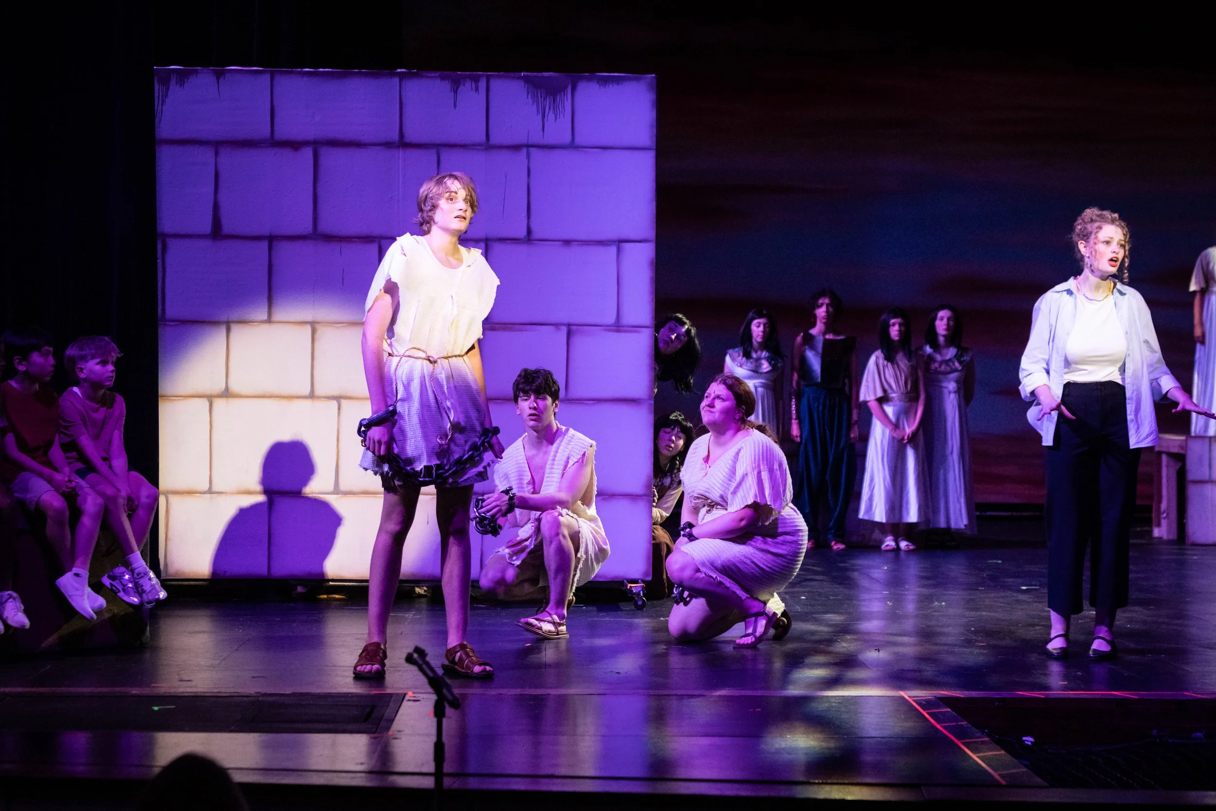 A theatrical stage scene with multiple actors in costume, some seated, some kneeling, and others standing, with a purple-lit cubic structure in the background.