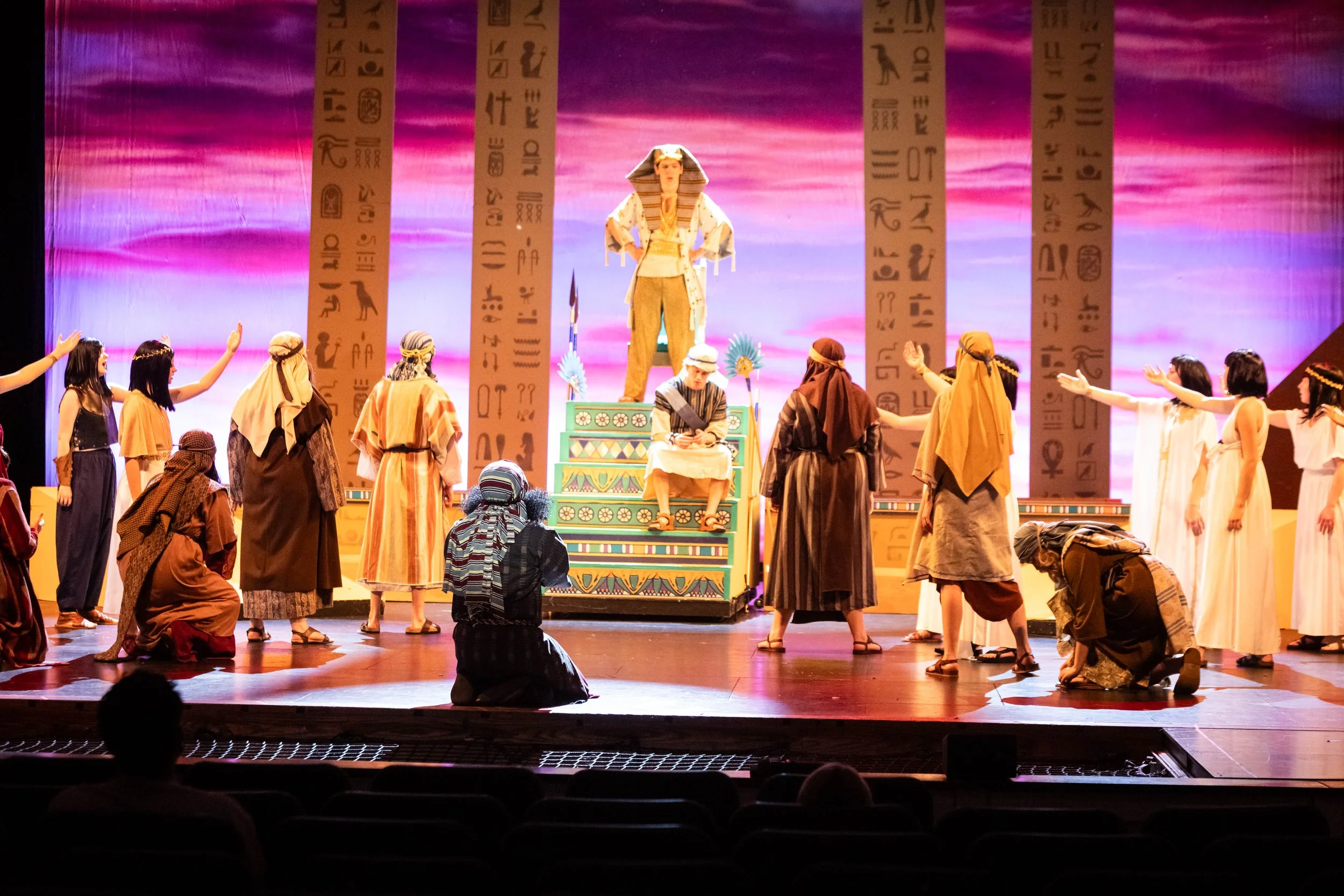 A theatrical performance on stage with actors in Egyptian costumes, some kneeling and others standing, with a backdrop featuring hieroglyphics and pink and purple lighting.