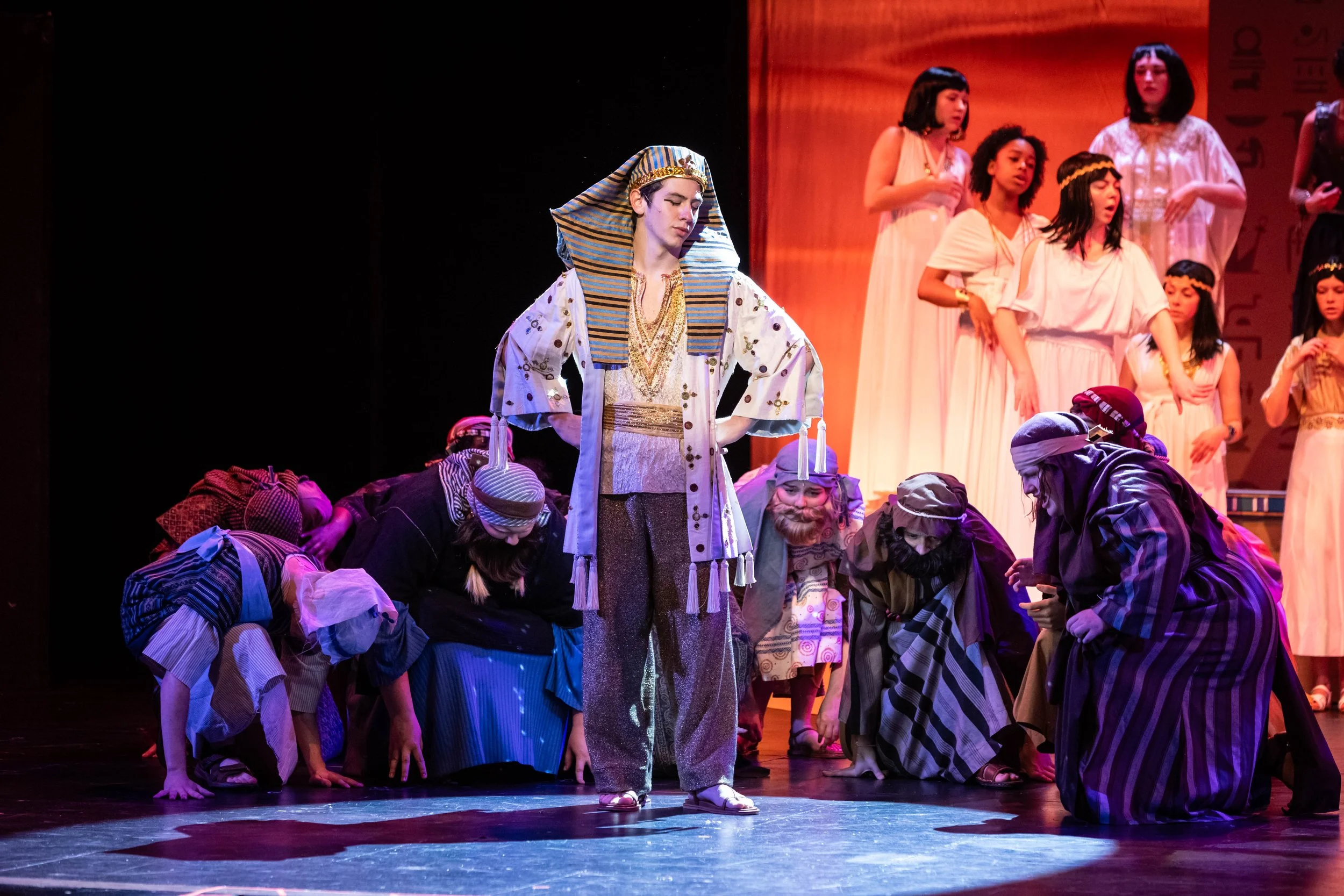 A theatrical stage scene with young performers dressed in costumes. The central figure is a young male dressed as an Egyptian pharaoh, wearing a striped headdress and ornate clothing, standing with hands on hips. Around him, others dressed as slaves 