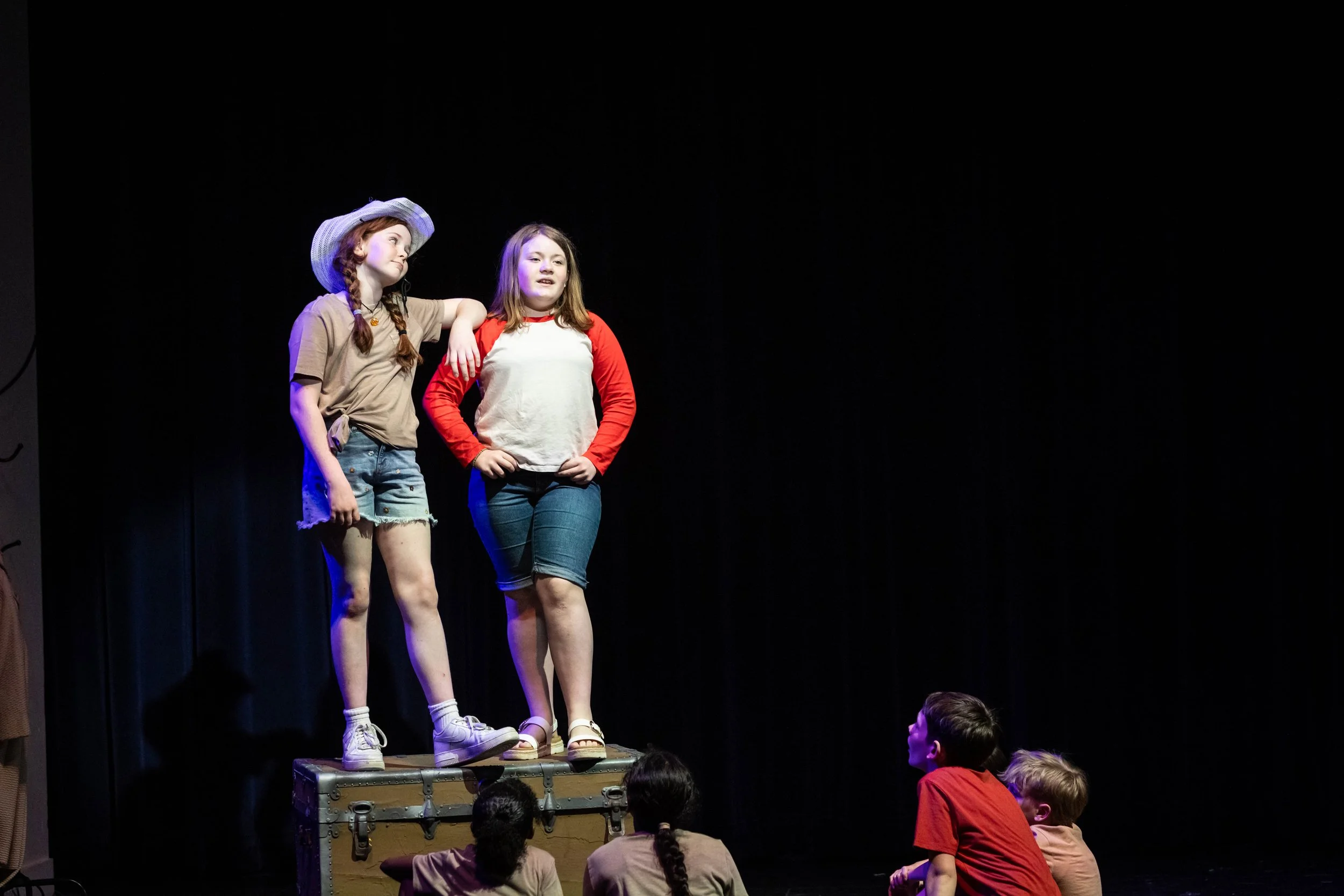 Two young girls stand on a platform with one girl’s arm around the other. They are performing on stage, with children watching in front of them. The girl on the left wears a cowboy hat, beige T-shirt, and shorts. The girl on the right wears a red and