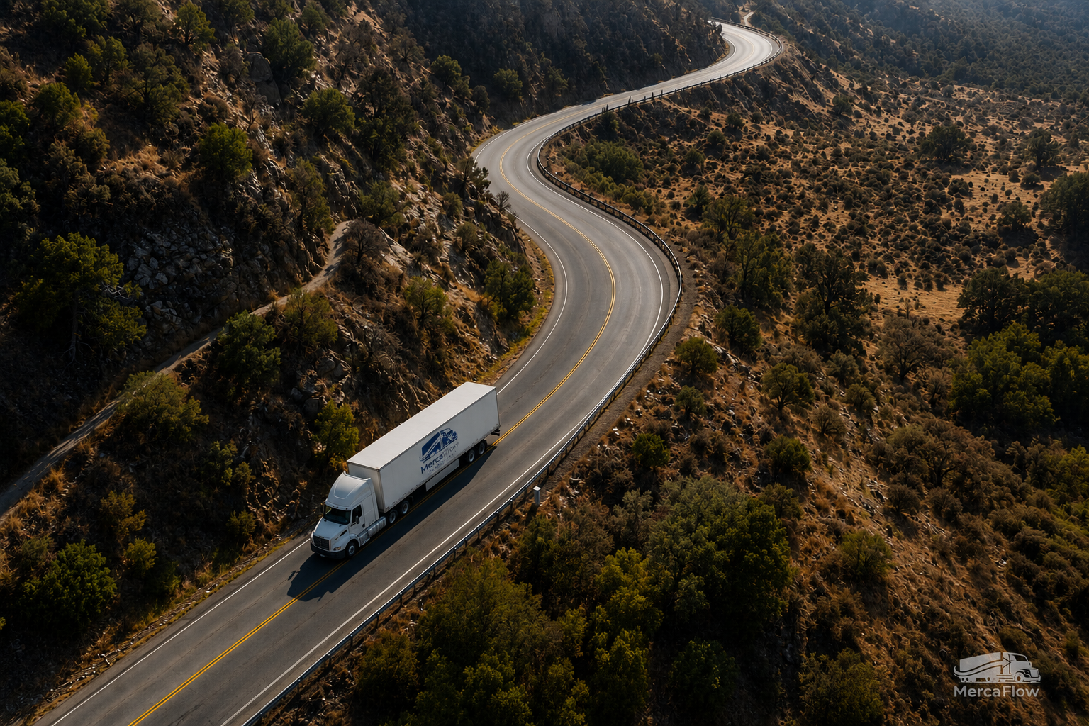 A white semi-truck driving on a winding mountain road surrounded by trees and rocky terrain.