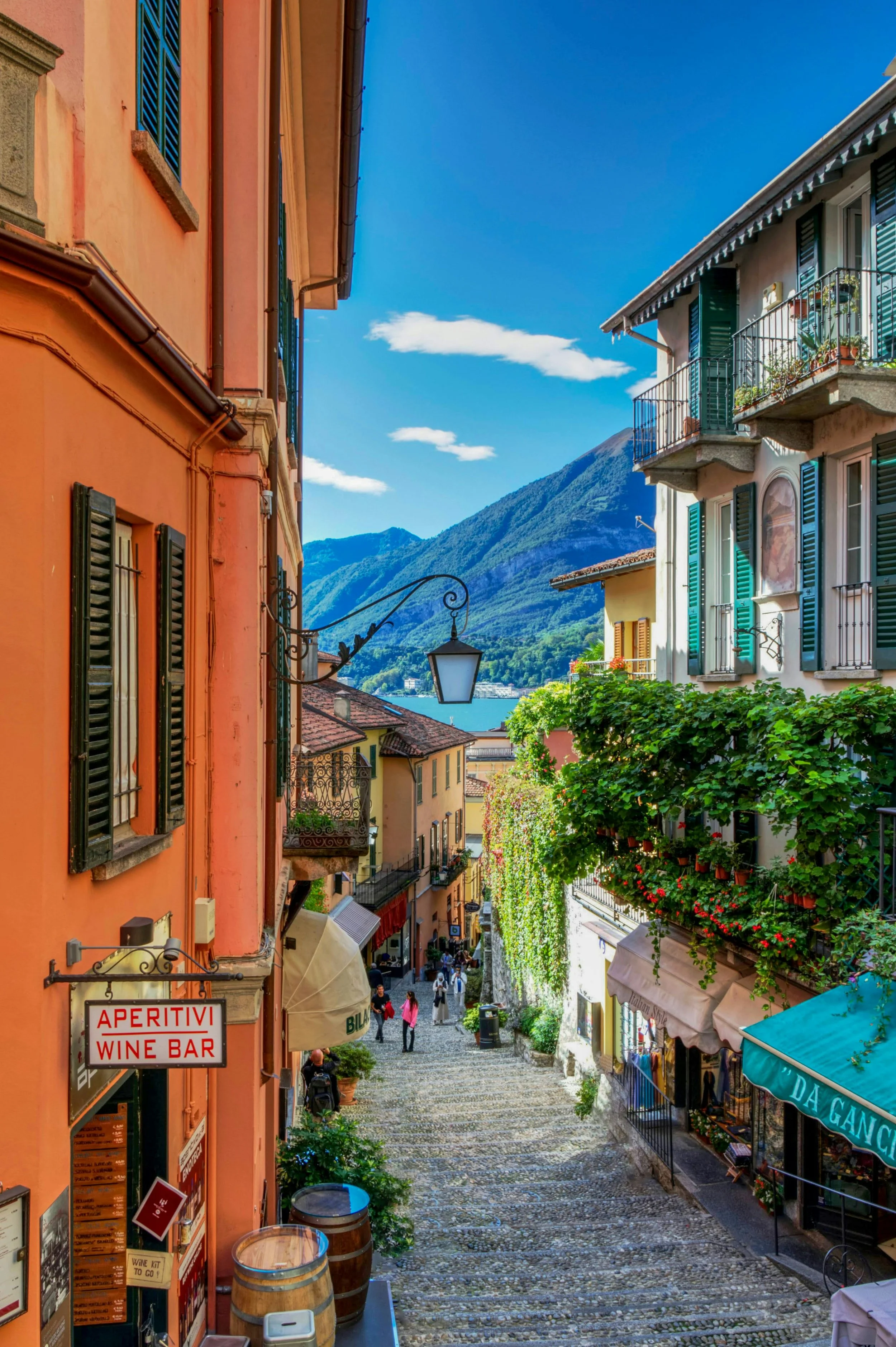 A cobblestone street lined with colorful Italian buildings with green shutters, balconies, and outdoor cafes, with a mountain in the background and a clear blue sky.