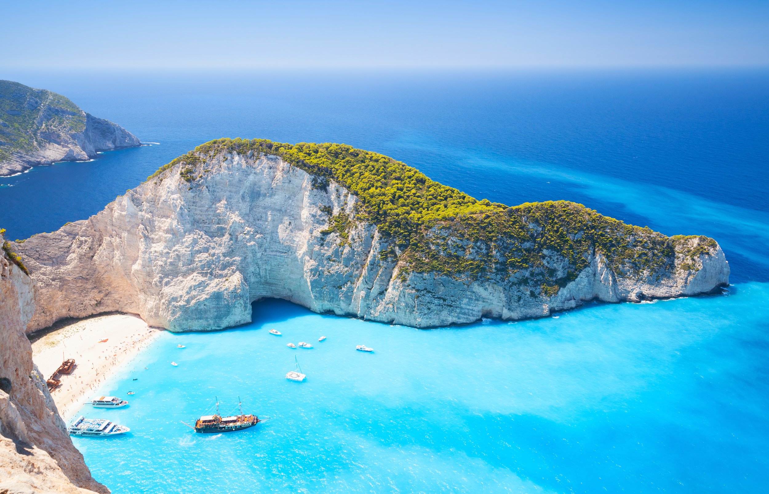 Cliffs with white rock formations overlooking the blue ocean with boats, and lush green vegetation on top of the cliffs.