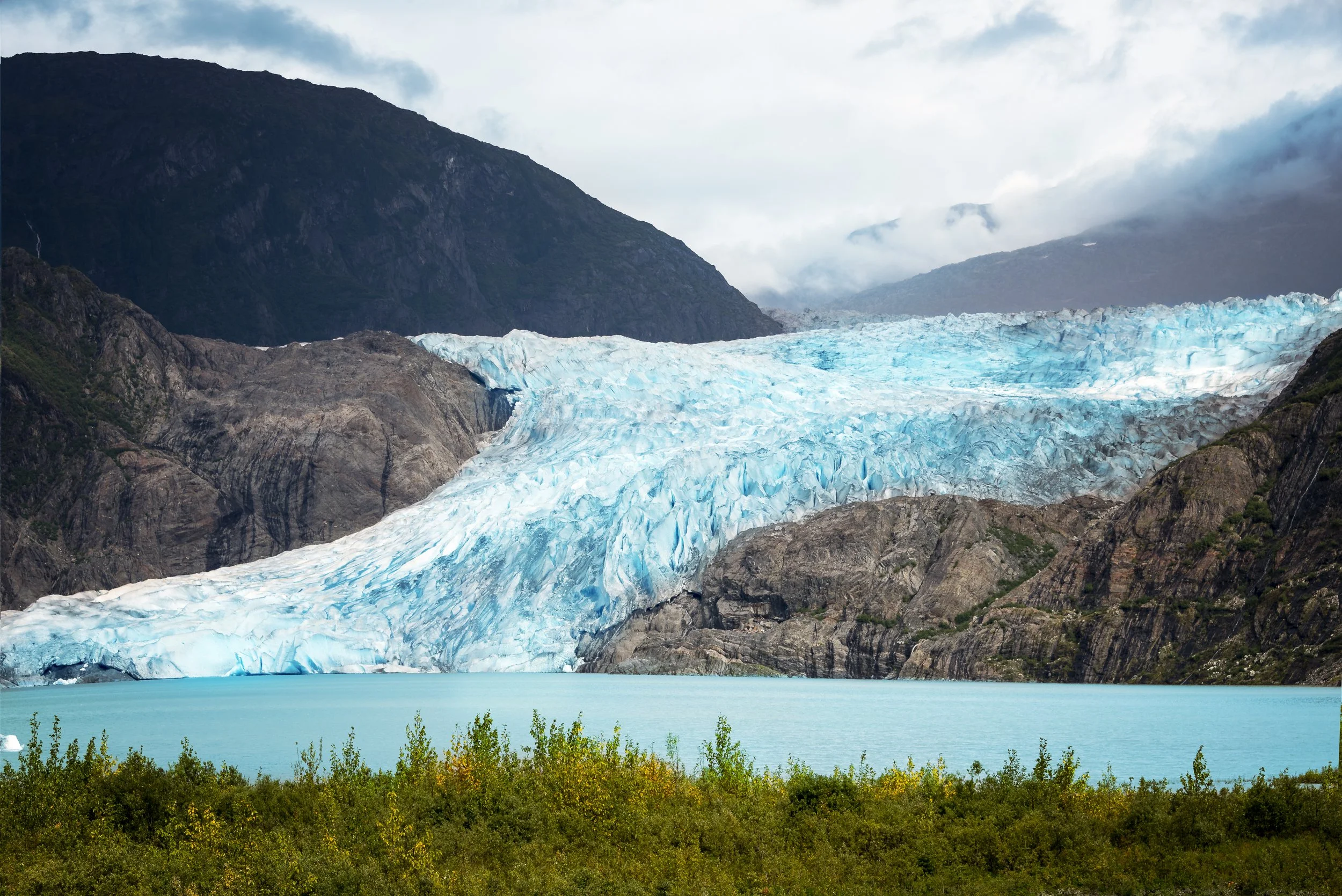 A glacier with blue ice flowing down between mountains, with a teal-colored lake in the foreground and green trees at the bottom.