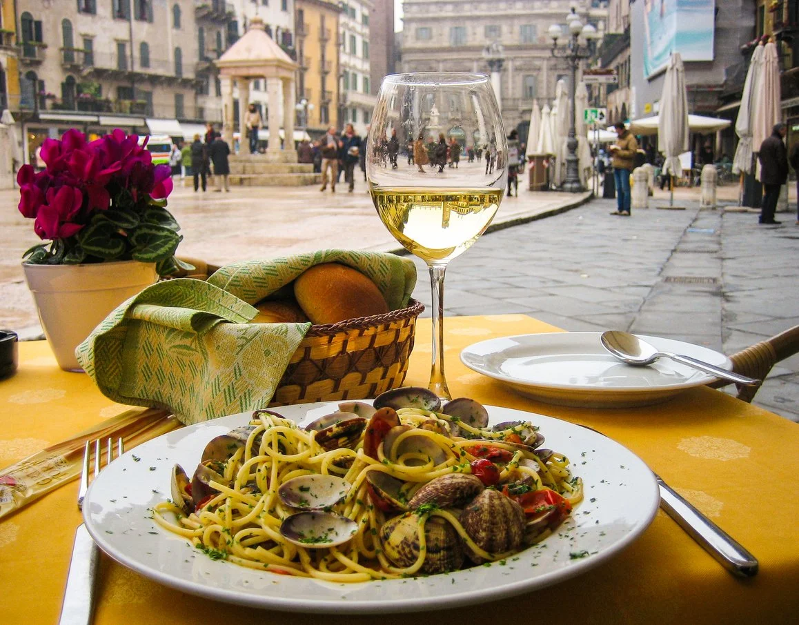 A plate of seafood pasta with clams, cherry tomatoes, and herbs on a table outside at a restaurant in a city square, with a glass of white wine, a basket of bread, and a potted plant nearby.
