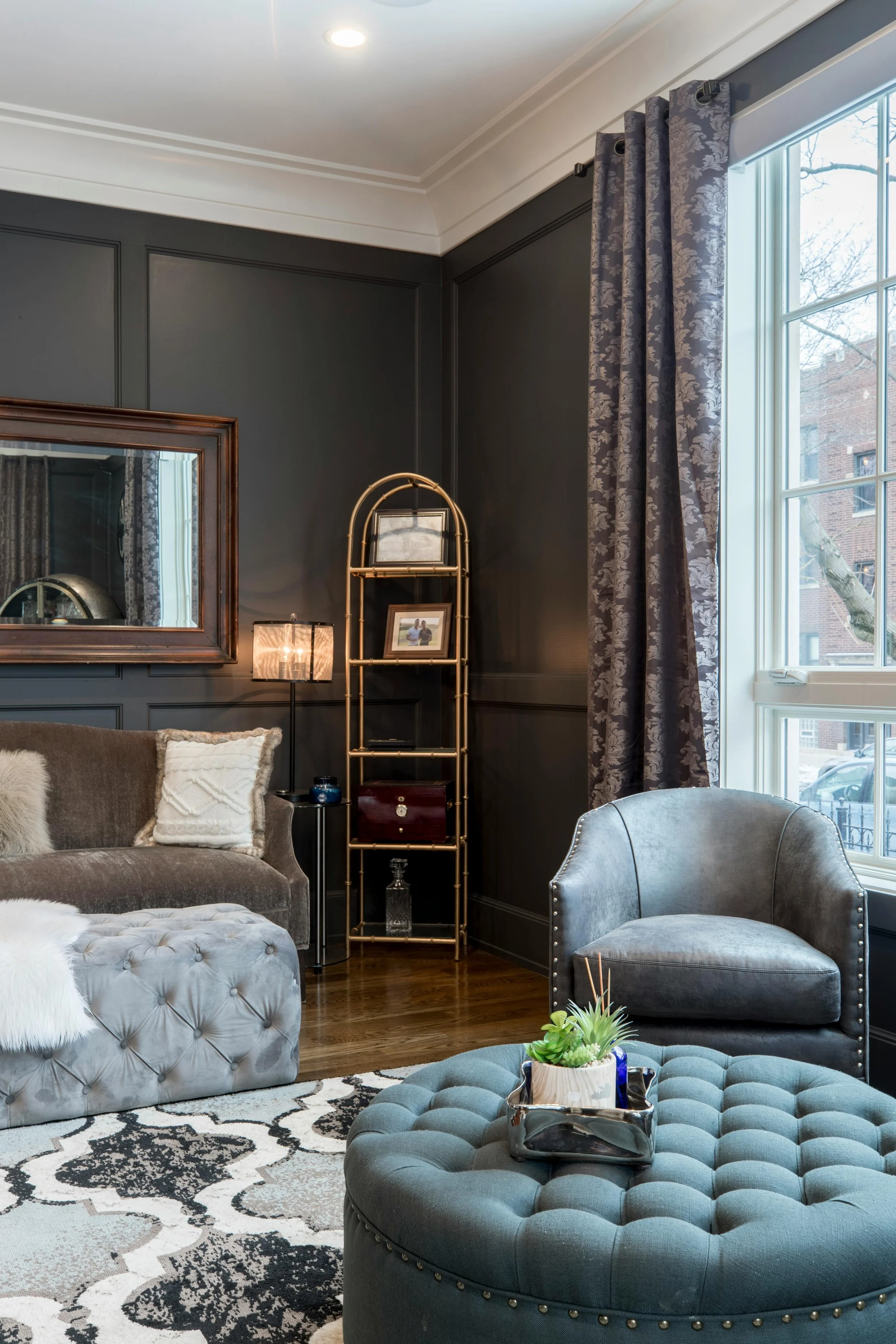 Elegant living room with dark gray walls, large window with patterned curtains, gray and plush furniture, gold shelving unit, mirror, table lamp, decorative pillows, area rug, and a small potted plant.