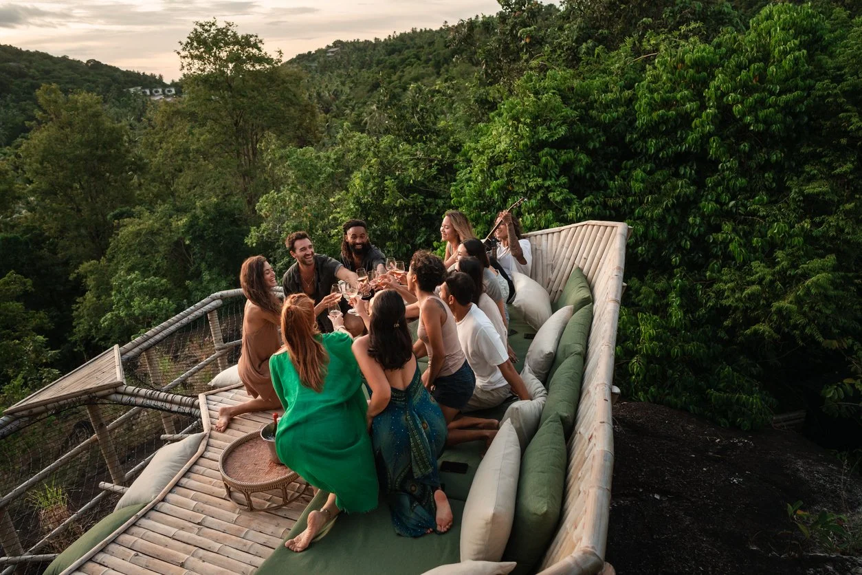 Group of people having a celebration or party on a rooftop terrace surrounded by lush green trees, during sunset.