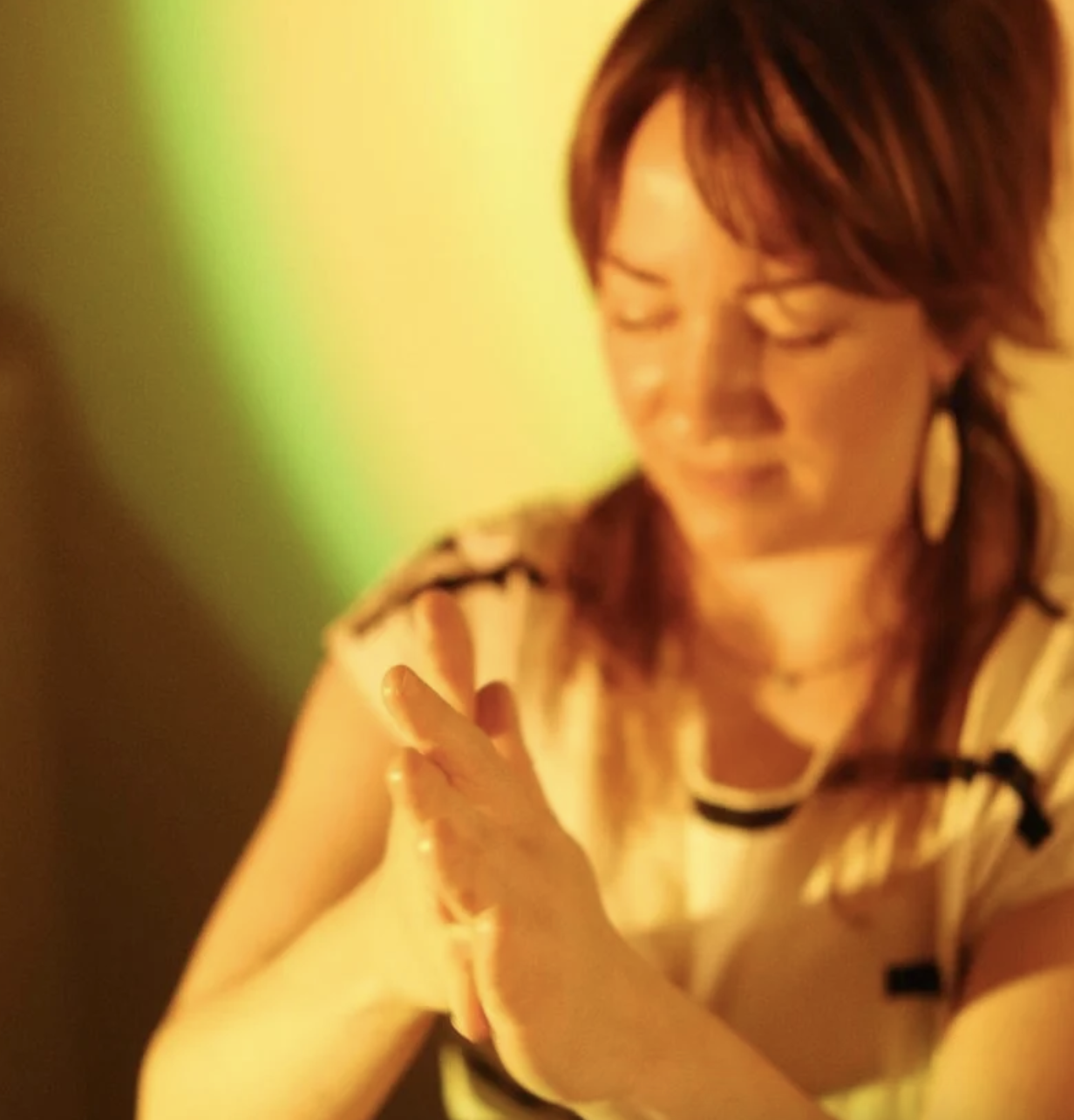 A woman with short brown hair smiling with her eyes closed, wearing a beige and black striped top, clasping her hands together, in a warmly lit setting with a blurred green background.