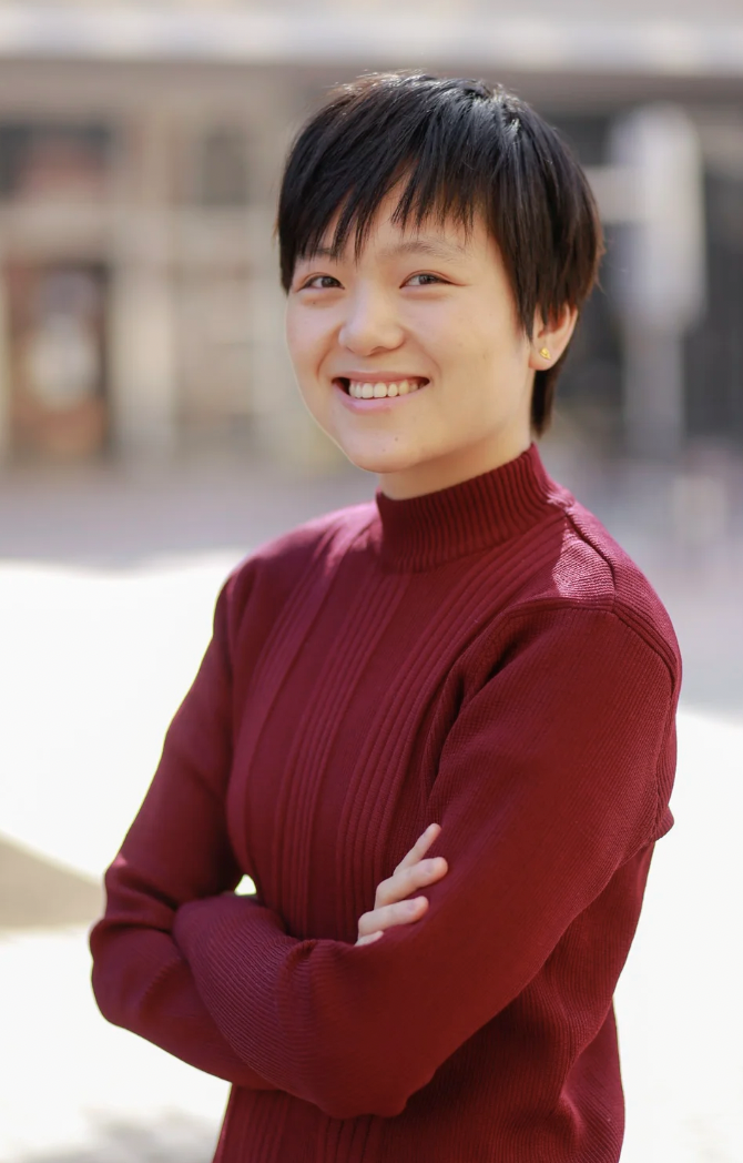 Smiling young woman with short black hair wearing a red sweater, standing outdoors with blurred background.