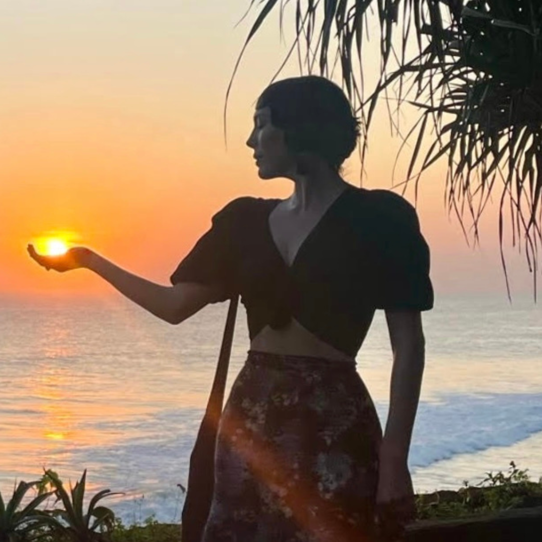 A woman standing on the beach at sunset, holding her hand in a cupped shape as if holding the setting sun, with ocean waves in the background and palm tree leaves overhead.