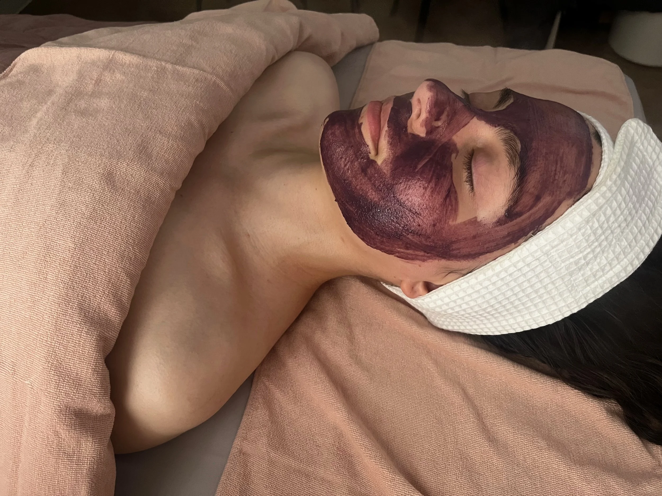 A woman lying on her back with her eyes closed, covered with a towel, with her face painted in dark reddish-brown facial mask.