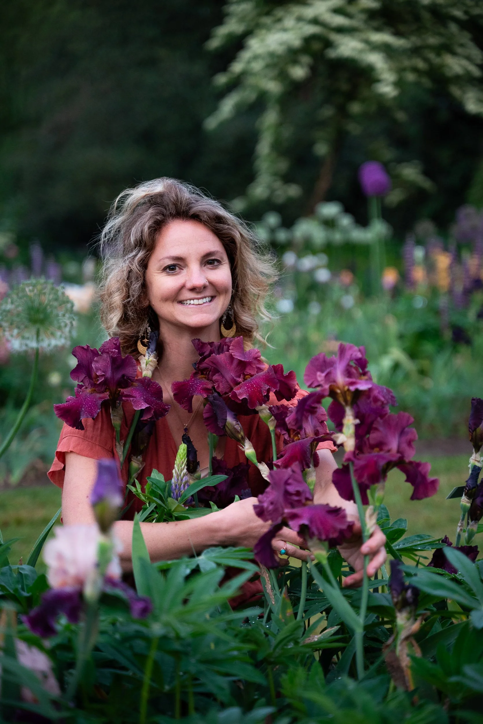 A woman with curly hair smiling while surrounded by purple flowers in a garden.