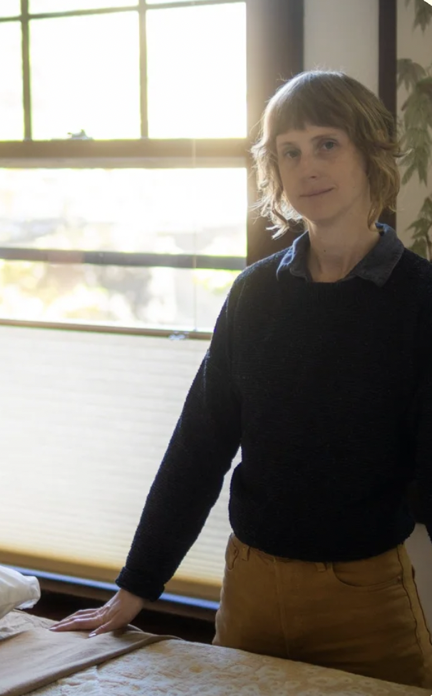 A person with short light brown hair standing indoors near a window with blinds, wearing a dark sweater over a collared shirt and tan pants, with a background of a wall and some greenery.