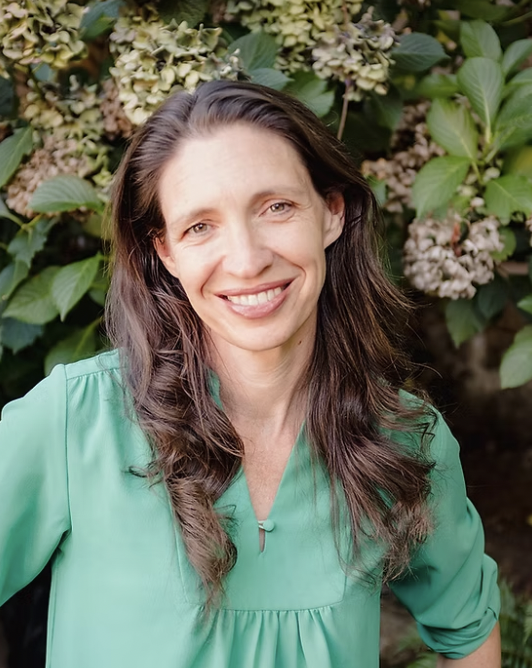 A woman with long brown hair wearing a green blouse, standing in front of a bush with green leaves and blooming flowers, smiling at the camera.