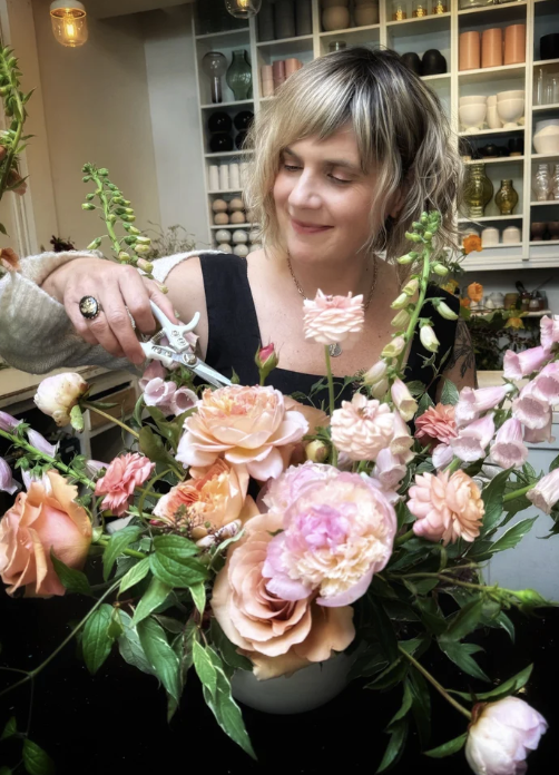 A woman with blond hair arranging a bouquet of pink and peach peonies in a floral shop.