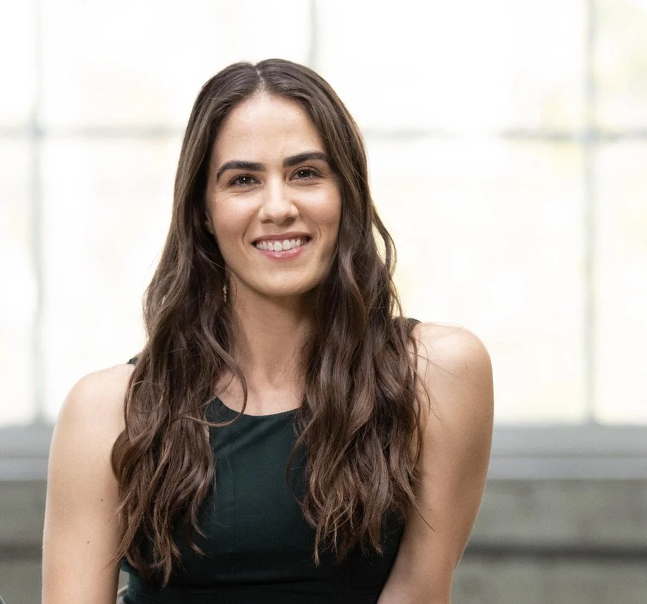 A young woman with long wavy brown hair, smiling, wearing a sleeveless black top.