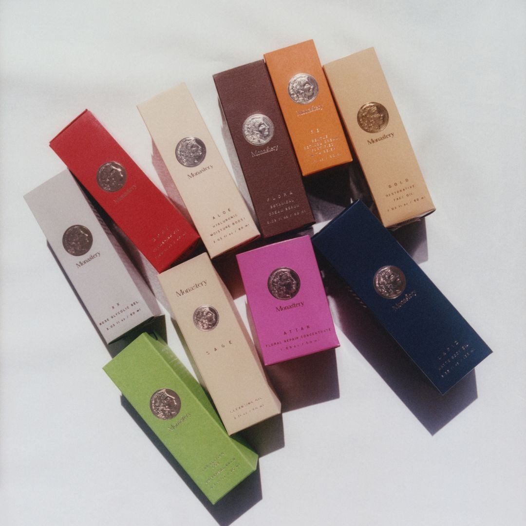 A collection of colorful perfume boxes arranged on a white surface, including red, beige, brown, orange, pink, navy blue, and green packages, each with a silver or gold emblem.