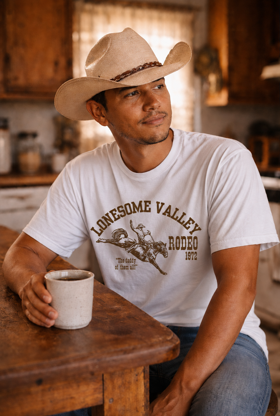 A man wearing a cowboy hat and a white t-shirt with a rodeo design sits at a wooden table holding a coffee mug.