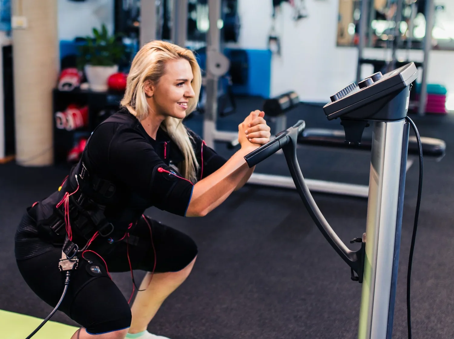 A woman exercising on a treadmill at the gym, smiling and wearing workout gear with monitoring sensors attached.